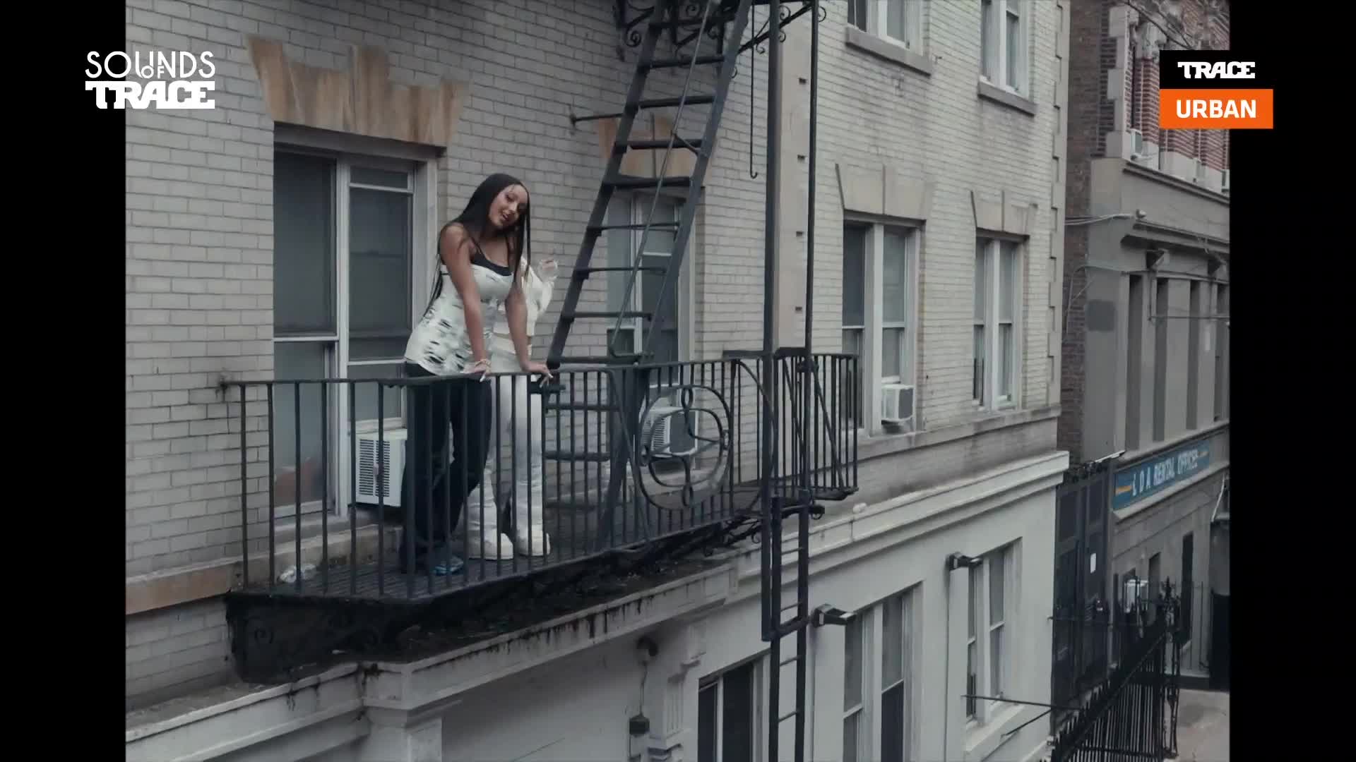 A young woman leans against the railing of a balcony, her gaze directed downwards. The scene, framed by the logos of Trace Urban and Sounds Trace, unfolds against a backdrop of brick buildings and a fire escape.
