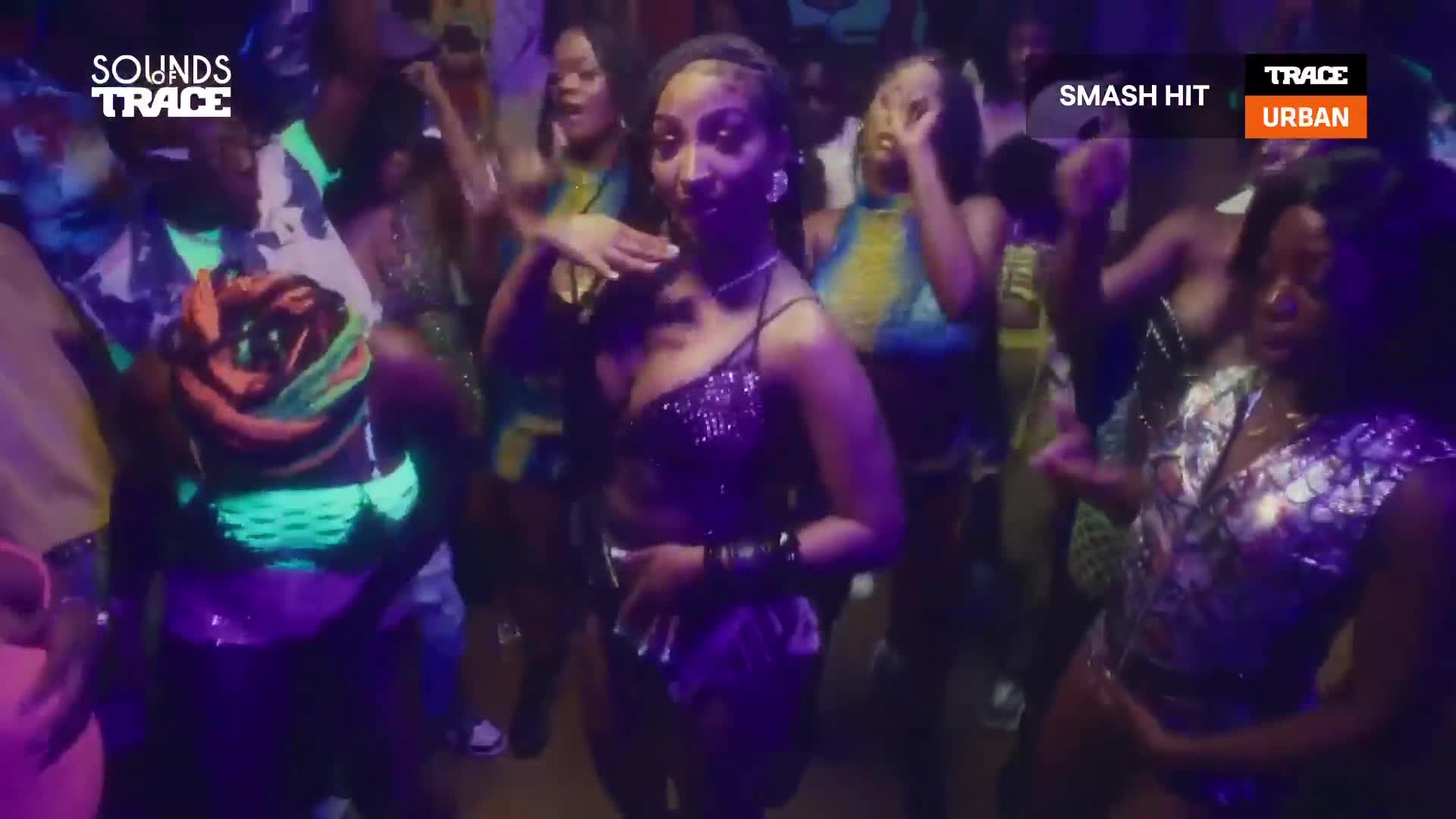 The crowd is moving to the music, with the woman in the center making a gesture with her hand. The club is lit with neon, and the Trace Urban logo is visible in the corner.
