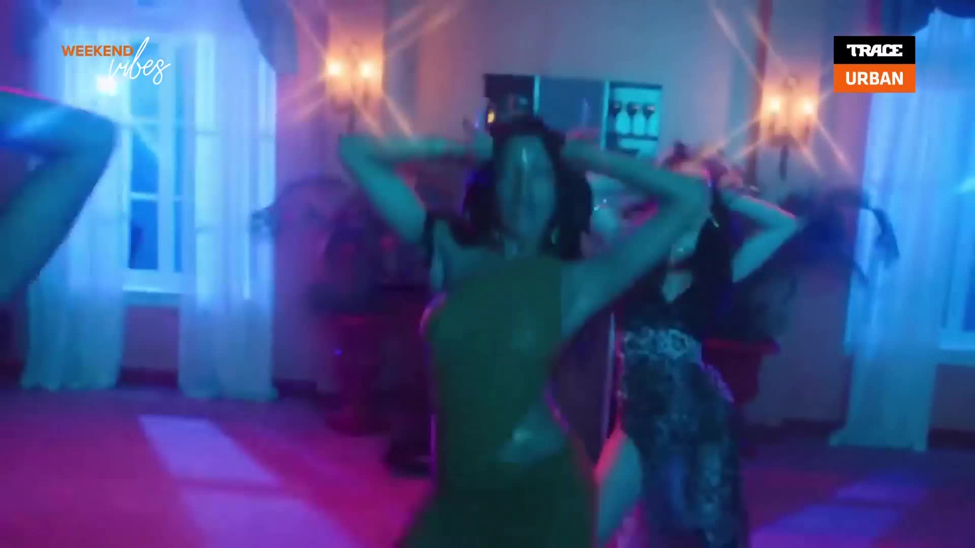 In a room bathed in blue light, a woman in a shiny dress moves with her arms raised above her head. Other figures, also dancing, are barely visible in the periphery.
