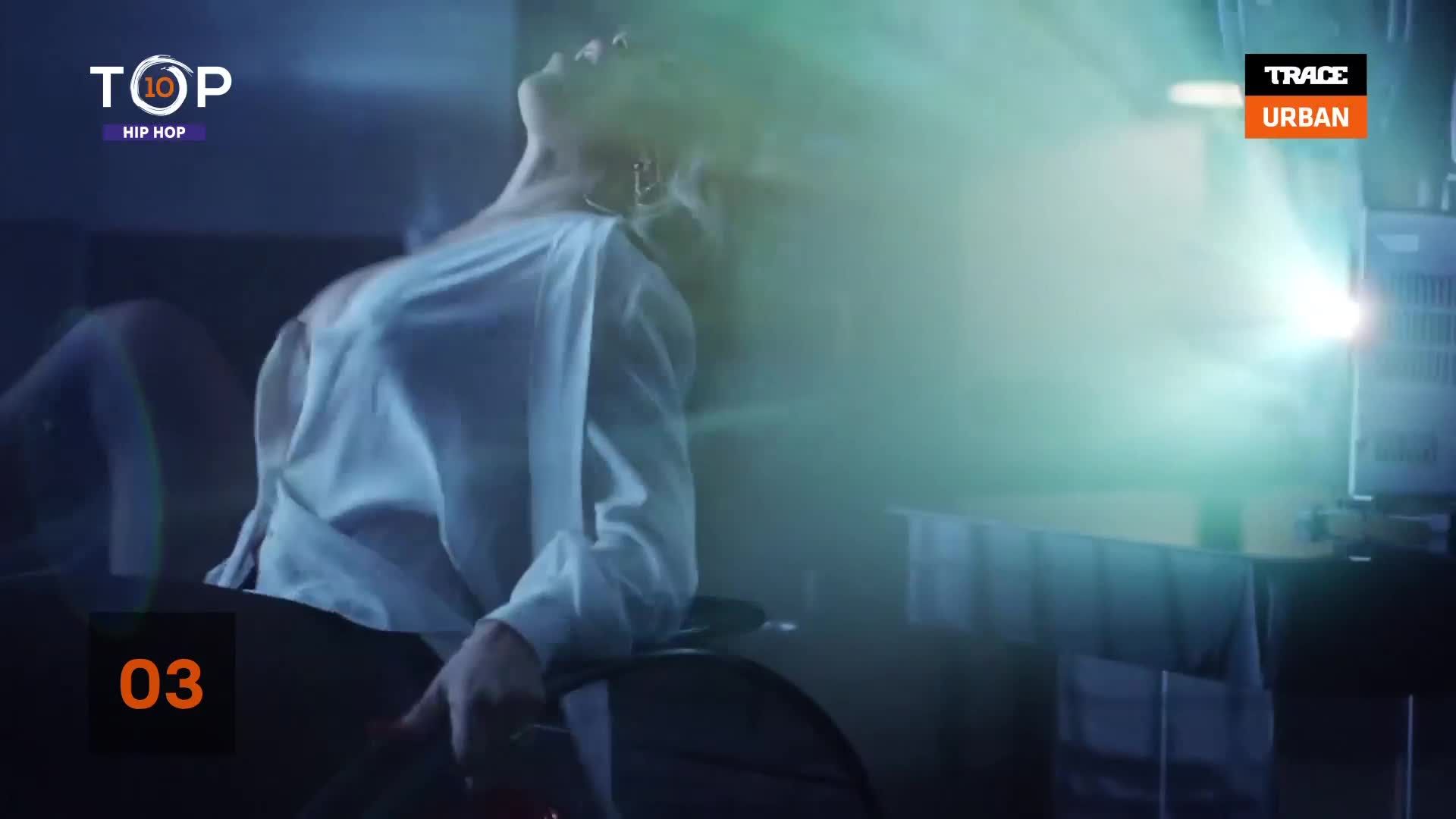 A woman in a white shirt leans back, her head tilted towards a bright projector beam. The scene, likely from a Trace Urban Top 10 Hip Hop countdown, is bathed in cool, blue light.
