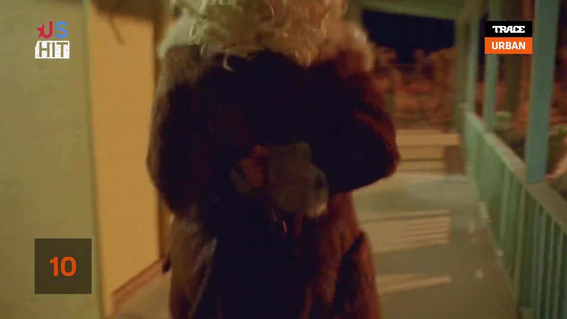 A person in a fur-trimmed coat is walking quickly, holding a small stuffed animal. The scene is lit by outdoor lights, with a porch railing visible to the right.
