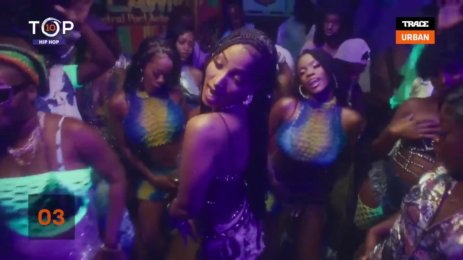 The woman in the center, in a dark dress, is turning towards the camera, her hair flowing. Dancers in matching blue and yellow tops move with her, all lit by the club's purple and green lights.
