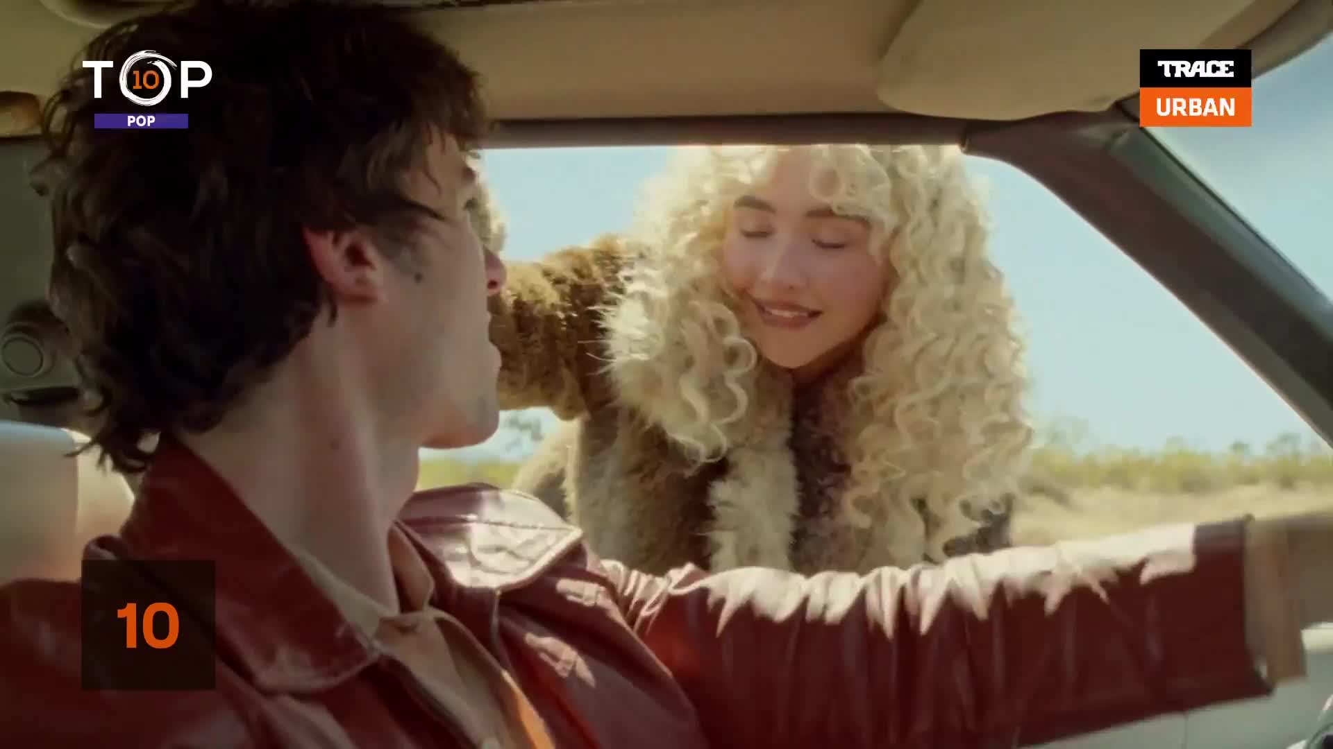 A woman with big, blonde curls leans into the car, smiling at the driver. He's turned toward her, wearing a leather jacket, as they cruise along a dusty road.
A woman with big, blonde curls leans into the car, smiling at the driver. He's turned toward her, wearing a leather jacket, as they cruise along a dusty road.