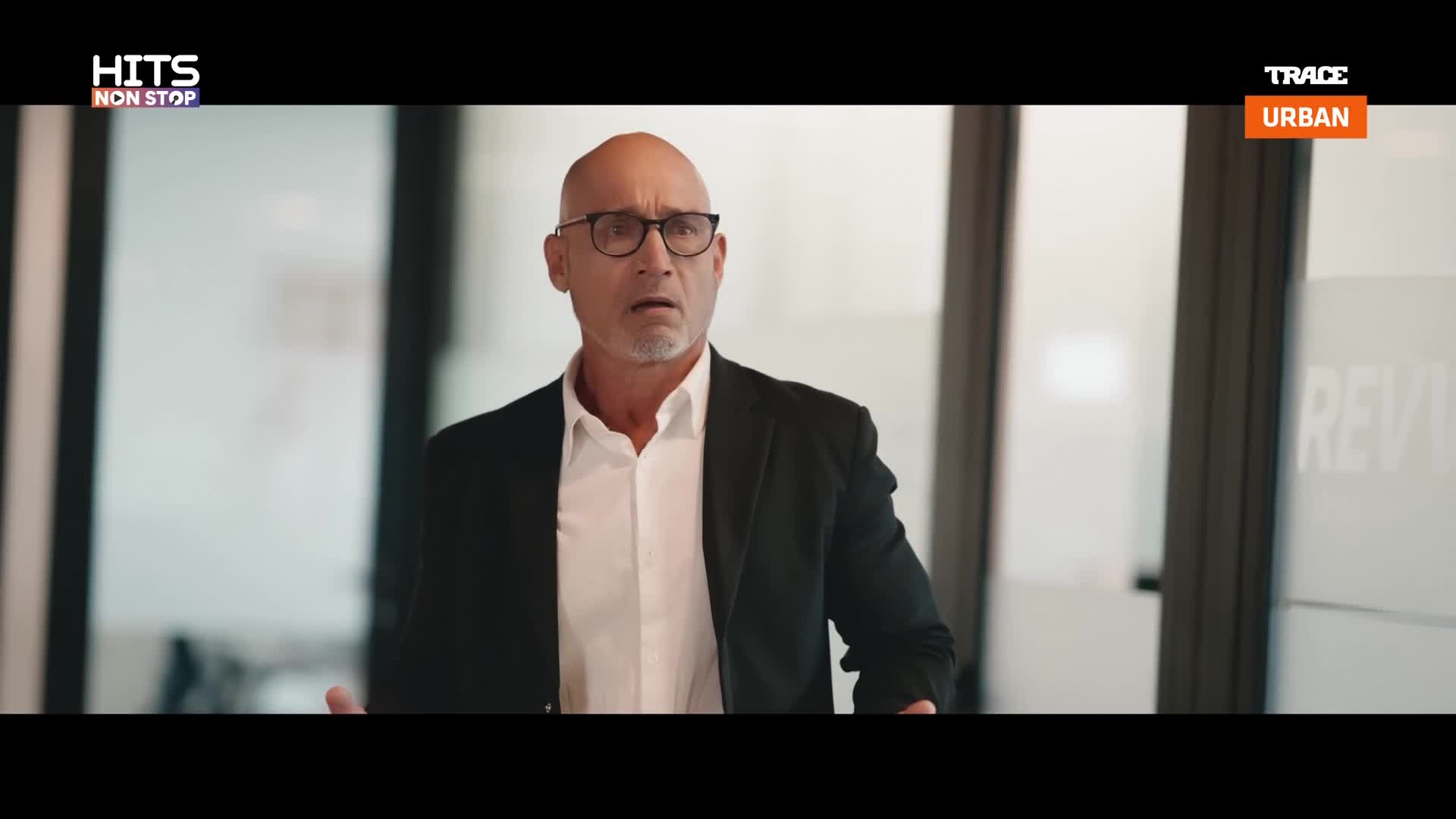 A man in a suit and glasses gestures with his hands, his face a mask of surprise. The Trace Urban logo flashes on screen, a vibrant orange against the muted background. A man in a suit and glasses gestures with his hands, his face a mask of surprise. The Trace Urban logo flashes on screen, a vibrant orange against the muted background.