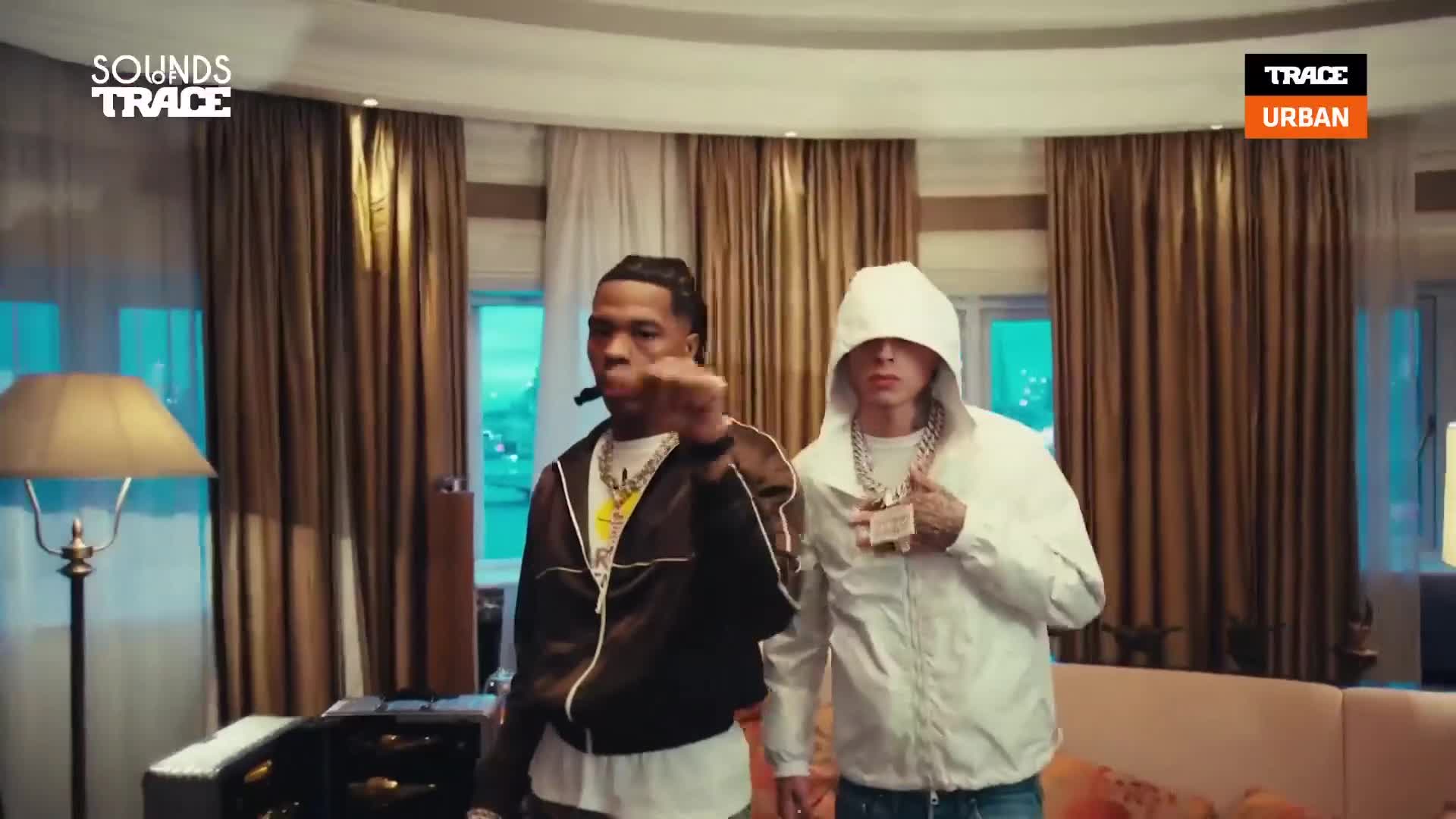 Two rappers stand in a room, one in a dark jacket and the other in a white hoodie. They're both wearing thick chains, looking directly at the camera as if ready for a performance. Two rappers stand in a room, one in a dark jacket and the other in a white hoodie. They're both wearing thick chains, looking directly at the camera as if ready for a performance.