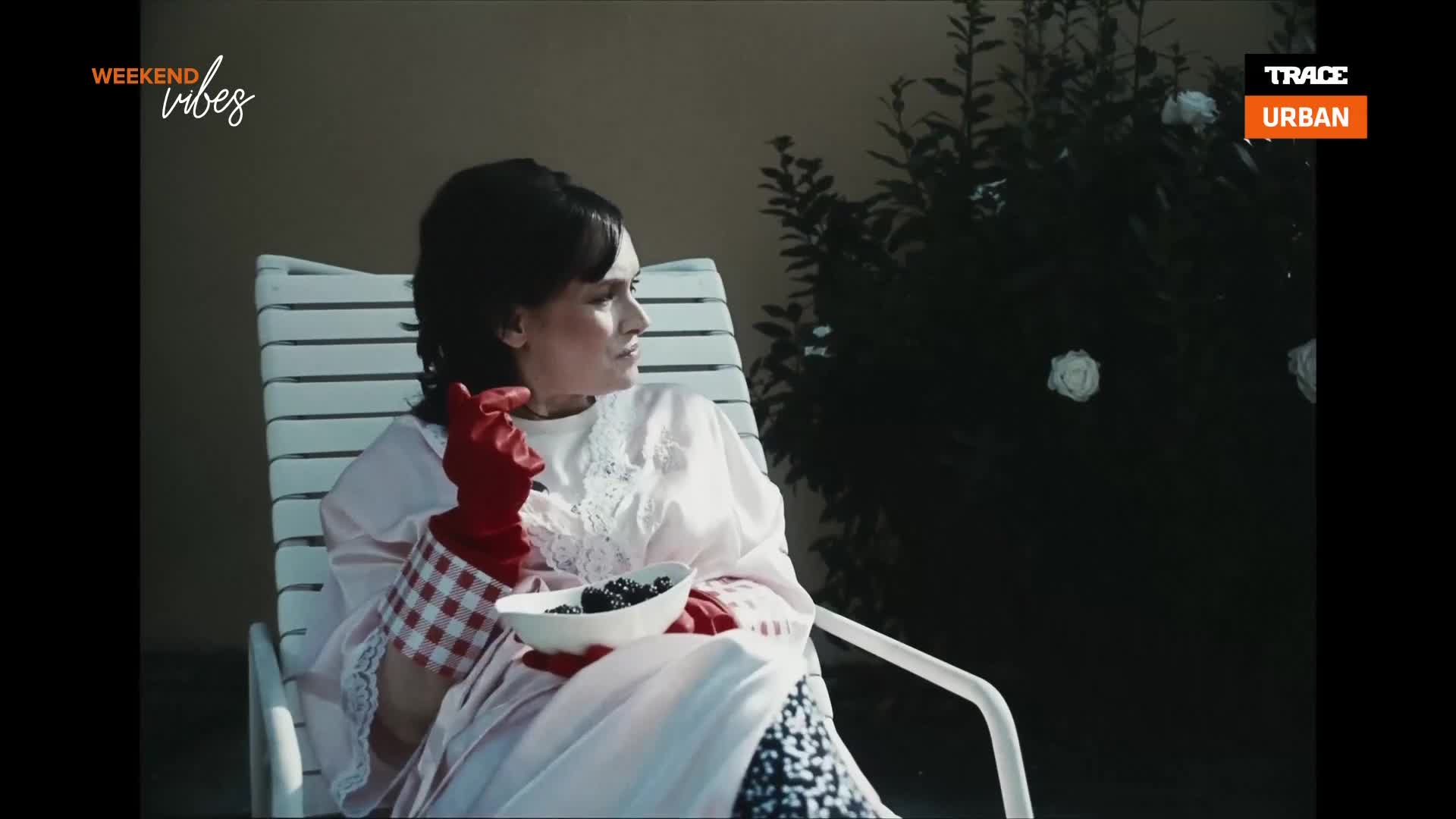 A woman in a pink dress and red gloves sits on a white lounge chair, holding a bowl of blackberries. She gazes off to the side, a faint smile on her face, as if lost in thought. A woman in a pink dress and red gloves sits on a white lounge chair, holding a bowl of blackberries. She gazes off to the side, a faint smile on her face, as if lost in thought.