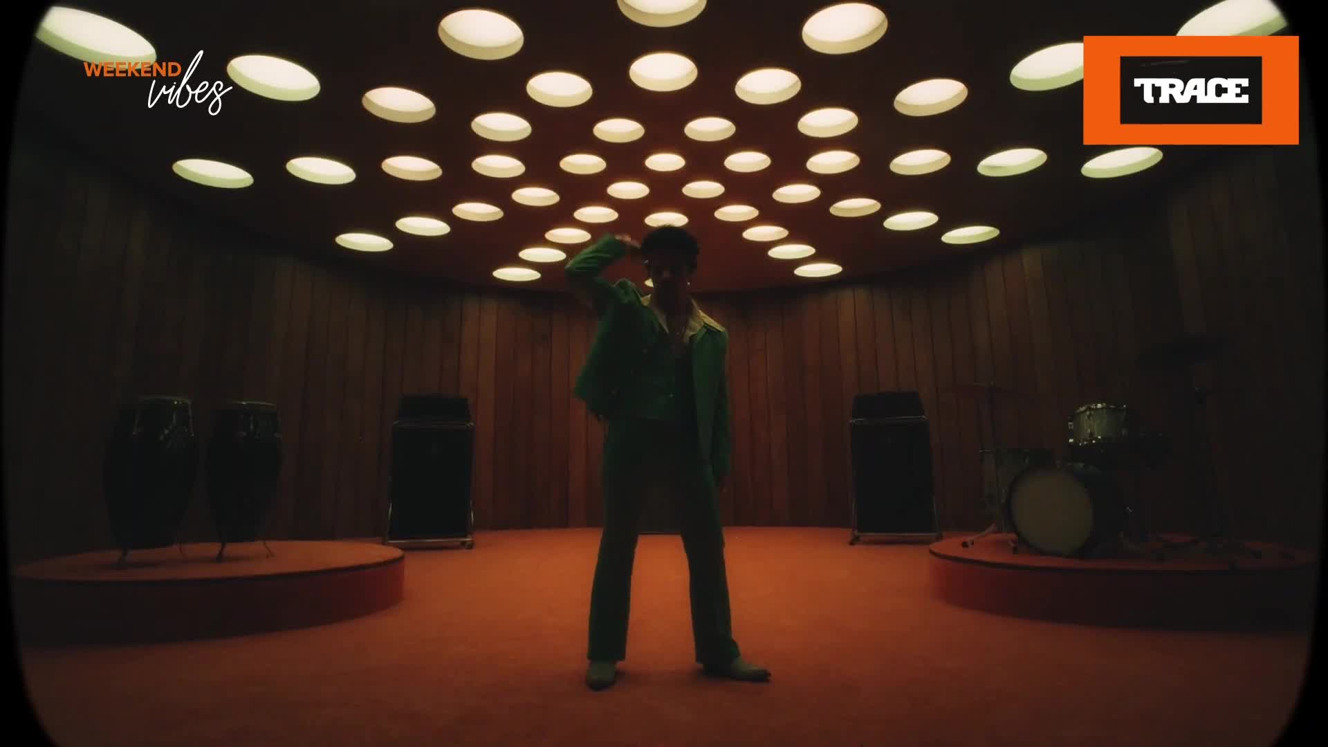 A lone figure in a green suit strikes a pose under a ceiling of circular lights. The room, with its wood-paneled walls and orange carpet, feels like a vintage French studio. A lone figure in a green suit strikes a pose under a ceiling of circular lights. The room, with its wood-paneled walls and orange carpet, feels like a vintage French studio.