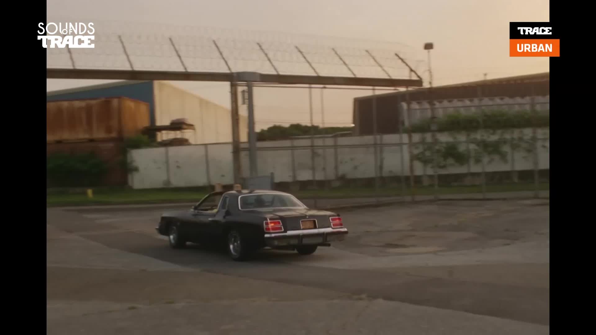 A dark, vintage coupe drifts around a corner, its tires kicking up dust. The low sun casts a warm glow on the industrial buildings behind it.