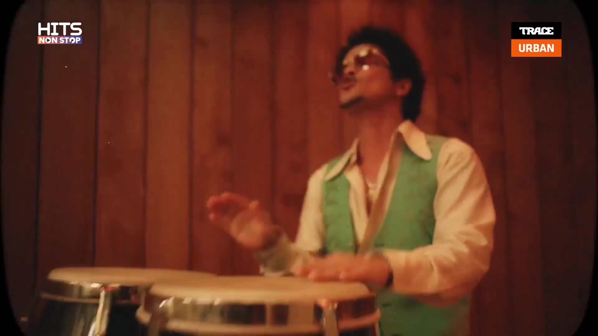 Hands move rhythmically over the conga drums, creating a beat. The performer, sporting a vintage vest, is lost in the music.