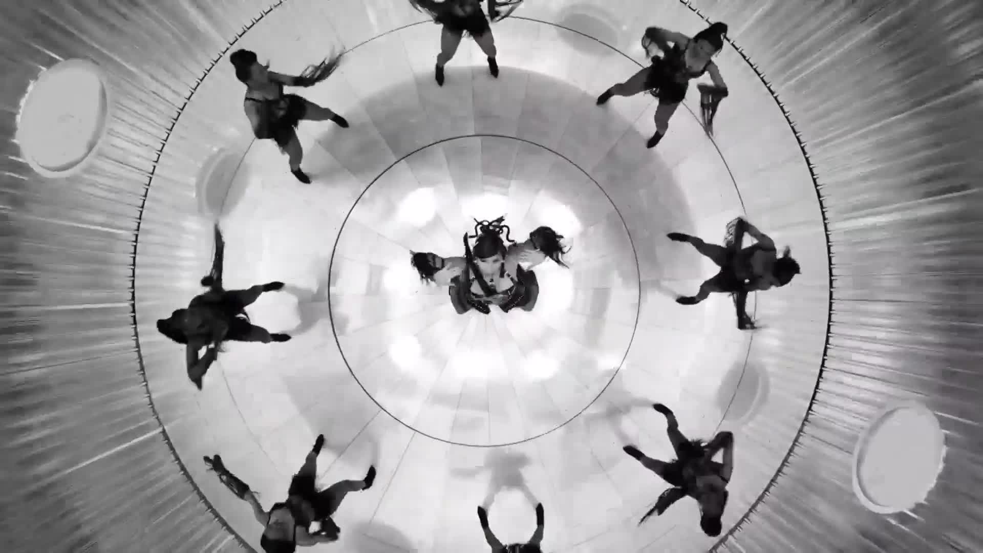 A central figure with elaborate hair stands with arms outstretched, surrounded by dancers in a circular formation. Their limbs are extended as they move in unison across the polished floor.