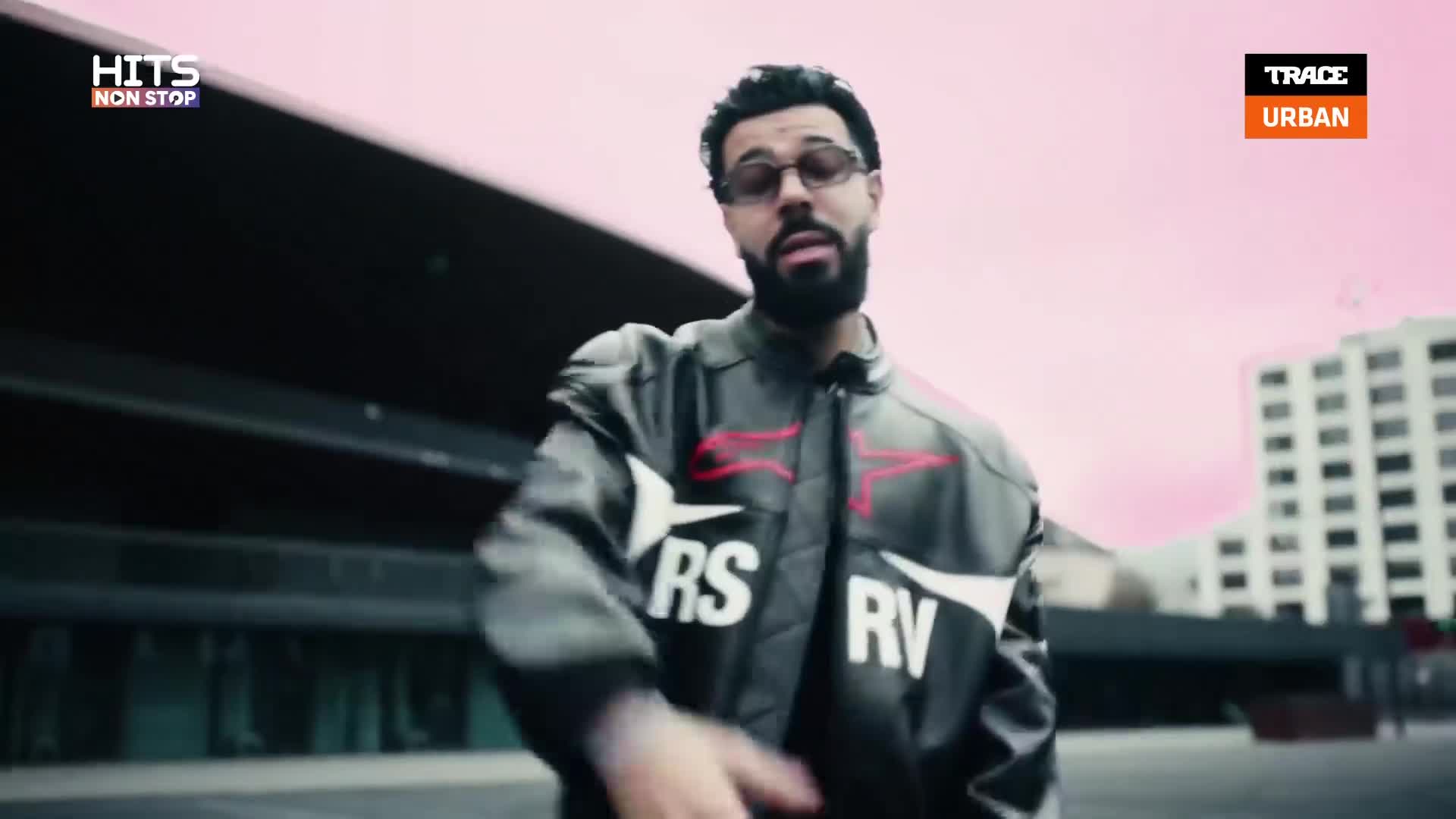 A man in a black jacket with white lettering moves his hands as he speaks. The pink sky behind him suggests a sunset over the city.