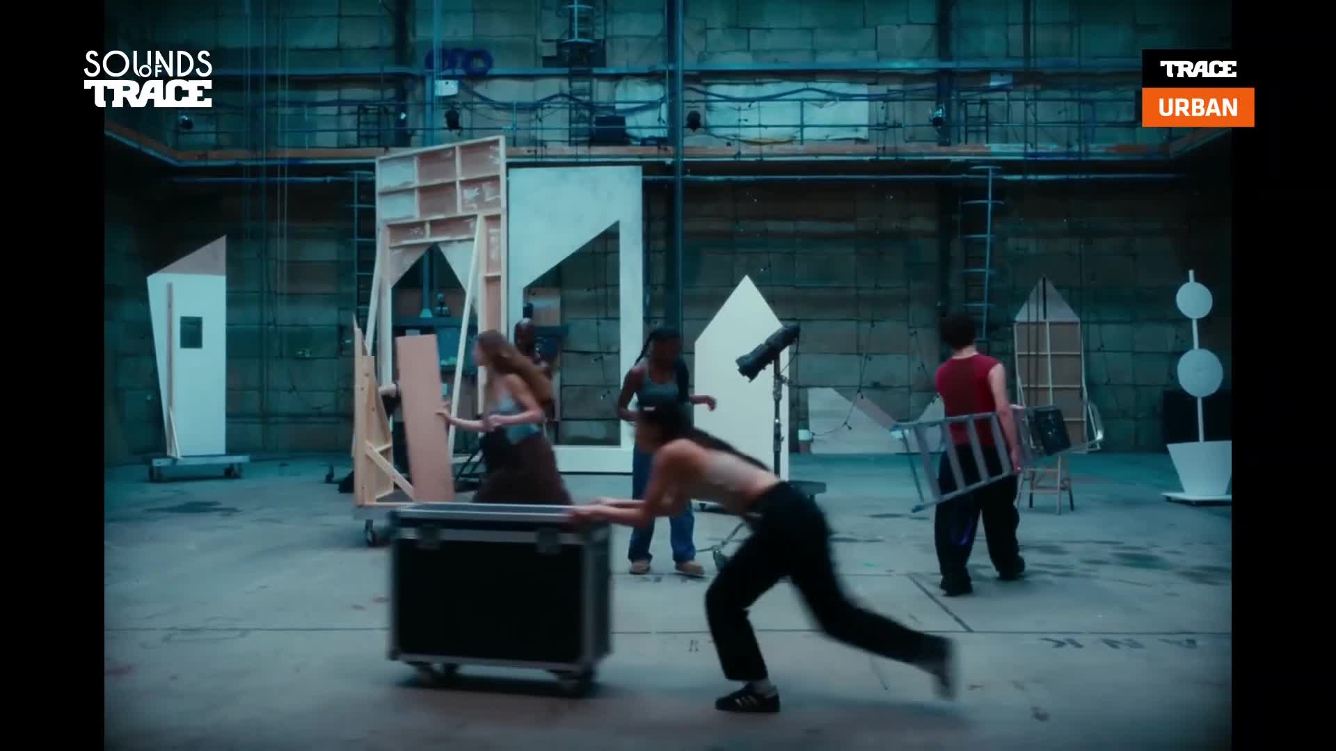 A woman in black pants lunges forward, pushing a large black case across the floor of this Parisian studio. Nearby, another woman in a brown skirt moves a large wooden panel, while a third person carries a metal ladder.