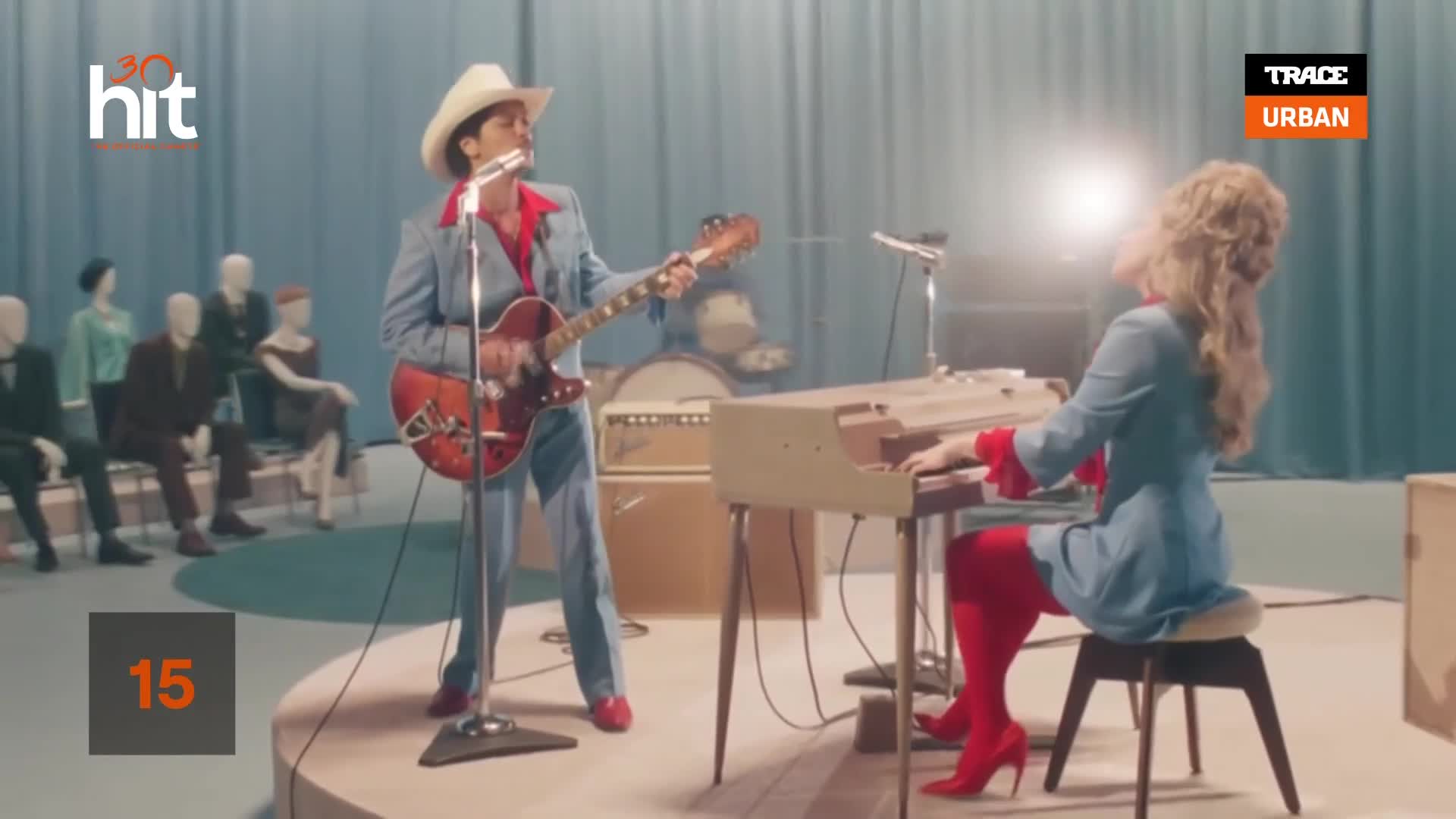 A man in a cowboy hat and a blue suit plays a red guitar while singing into a microphone. To his right, a woman with blonde hair plays a keyboard, her red boots catching the light.