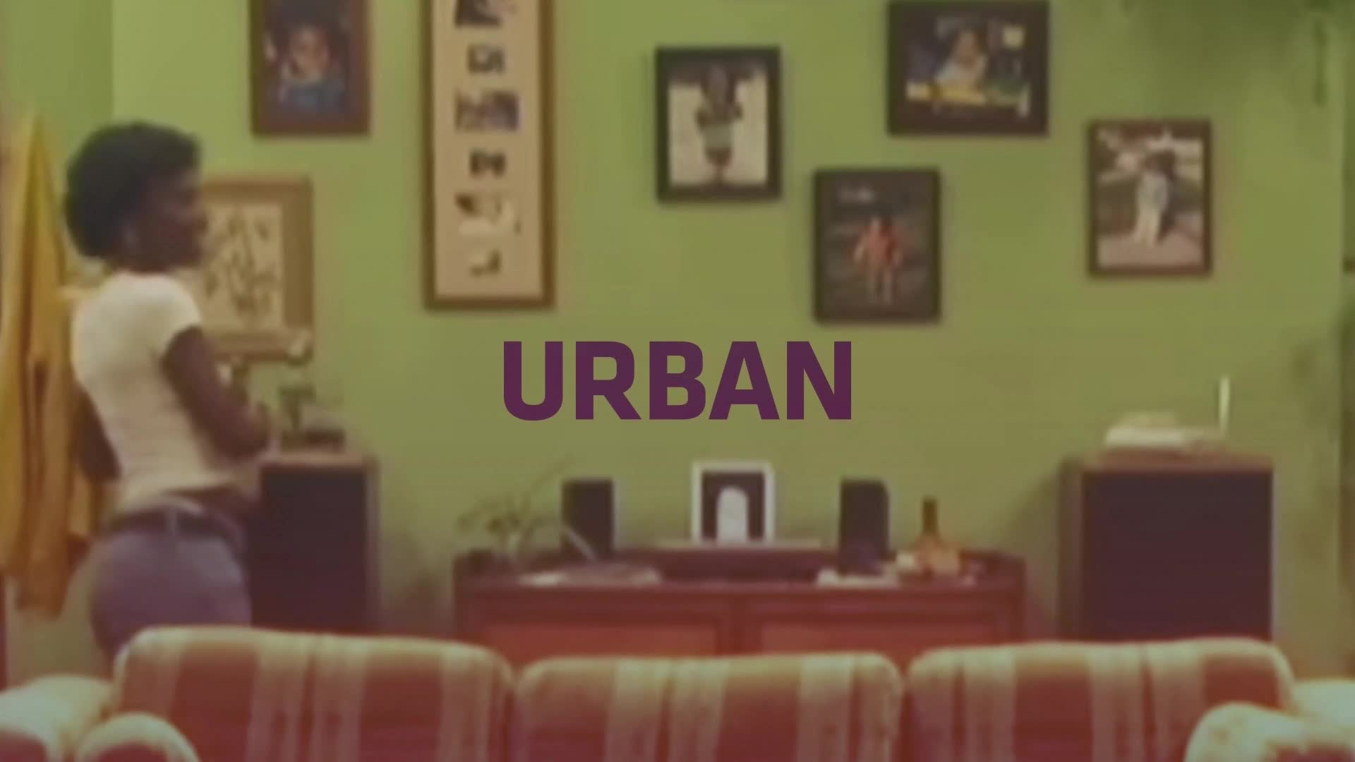 A young woman in a white t-shirt and jeans turns her head, her dark hair framing her face. The word "URBAN" is emblazoned across the pale green wall behind her.