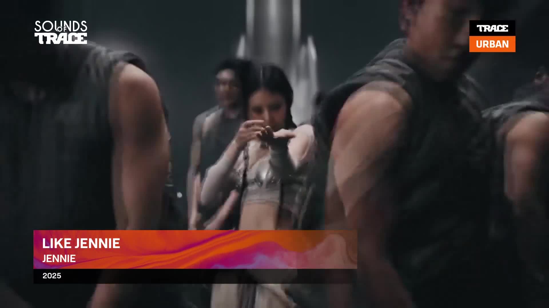 The camera pans across a group of dancers, their movements sharp and synchronized. In the center, a woman with dark hair holds a small object close to her face, her expression focused. The camera pans across a group of dancers, their movements sharp and synchronized. In the center, a woman with dark hair holds a small object close to her face, her expression focused.
