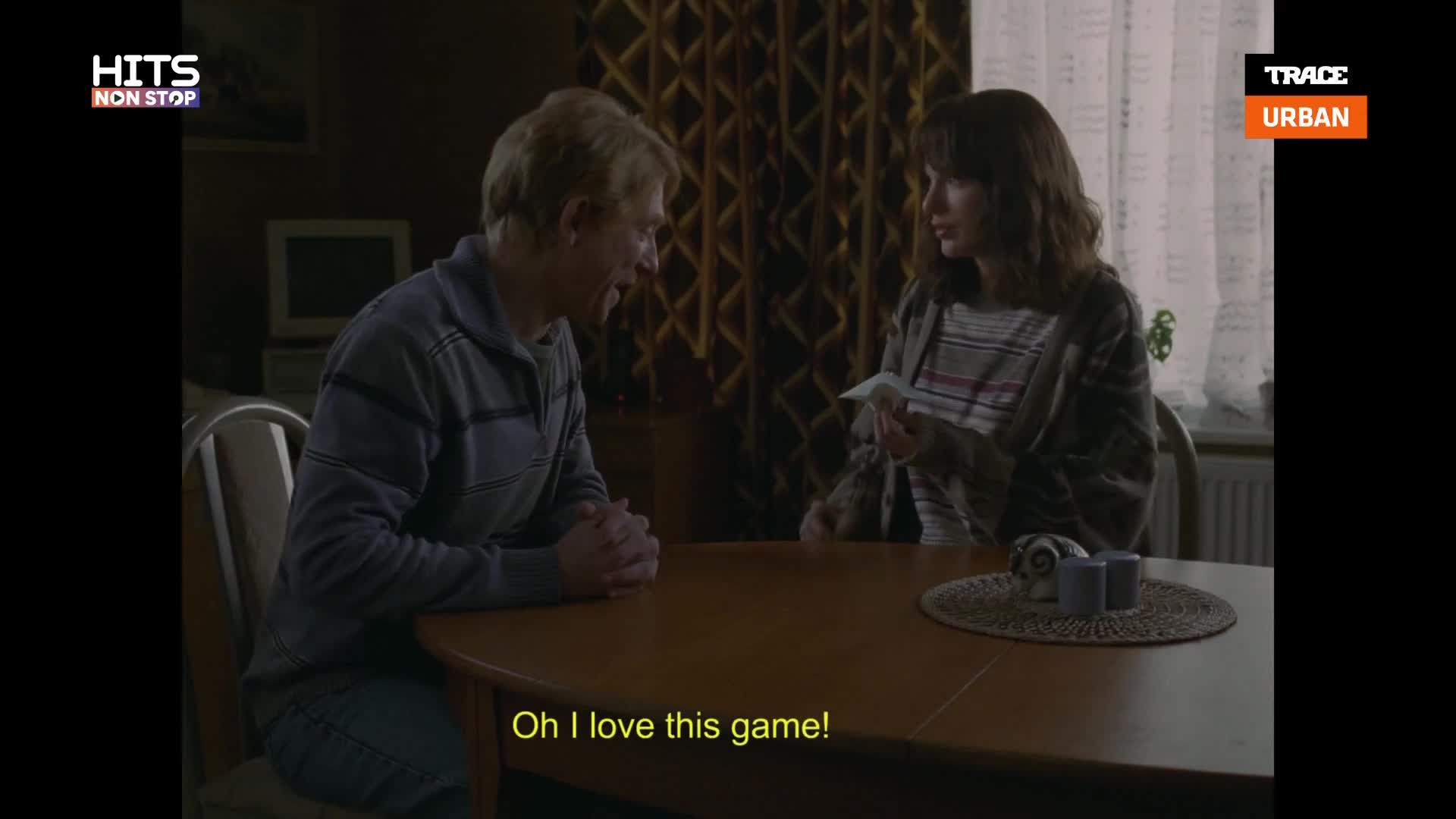 A man with blond hair leans forward, hands clasped, as a woman across the table holds a small paper. The woman, with dark, wavy hair, looks intently at the paper she holds. A man with blond hair leans forward, hands clasped, as a woman across the table holds a small paper. The woman, with dark, wavy hair, looks intently at the paper she holds.