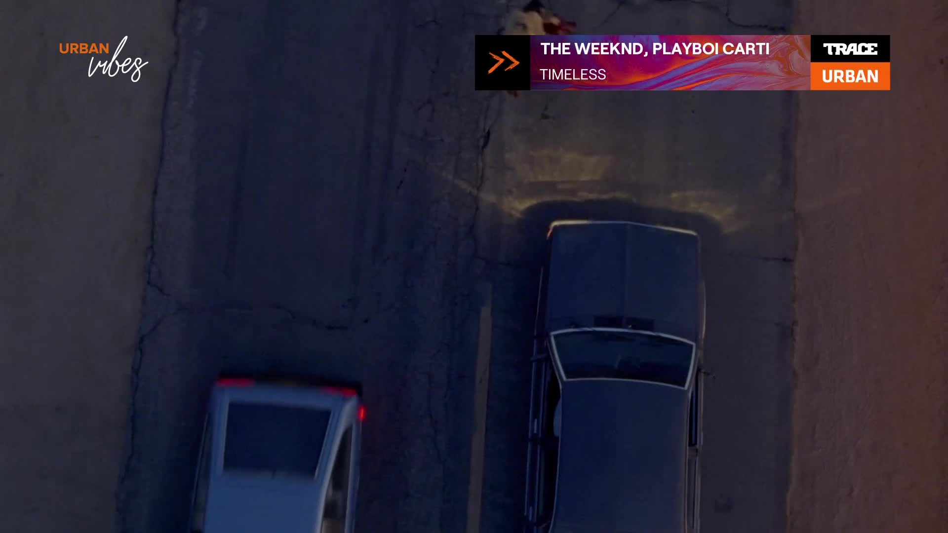 Two cars drive down a dark road, their headlights cutting through the night. A graphic overlay announces "The Weeknd, Playboi Carti" and "Timeless" from Trace Urban. Two cars drive down a dark road, their headlights cutting through the night. A graphic overlay announces "The Weeknd, Playboi Carti" and "Timeless" from Trace Urban.