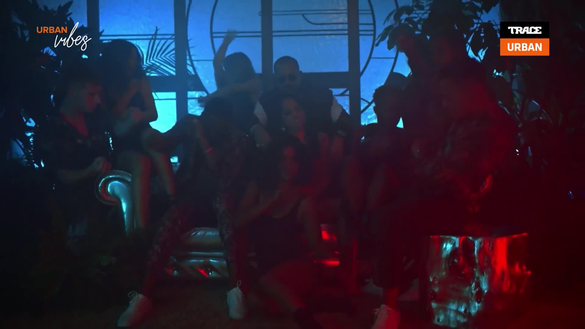 A group of people are dancing and moving in a dimly lit space, illuminated by blue and red lights. The Trace Urban logo appears in the corner, suggesting a music video or performance. A group of people are dancing and moving in a dimly lit space, illuminated by blue and red lights. The Trace Urban logo appears in the corner, suggesting a music video or performance.
