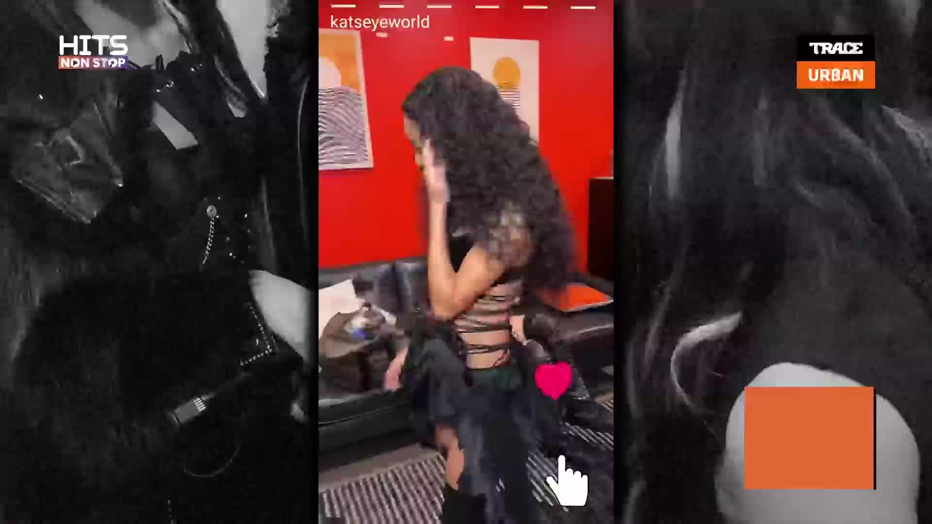 A woman with long, dark, curly hair is dancing in a black outfit. She's in a room with red walls and a black couch.