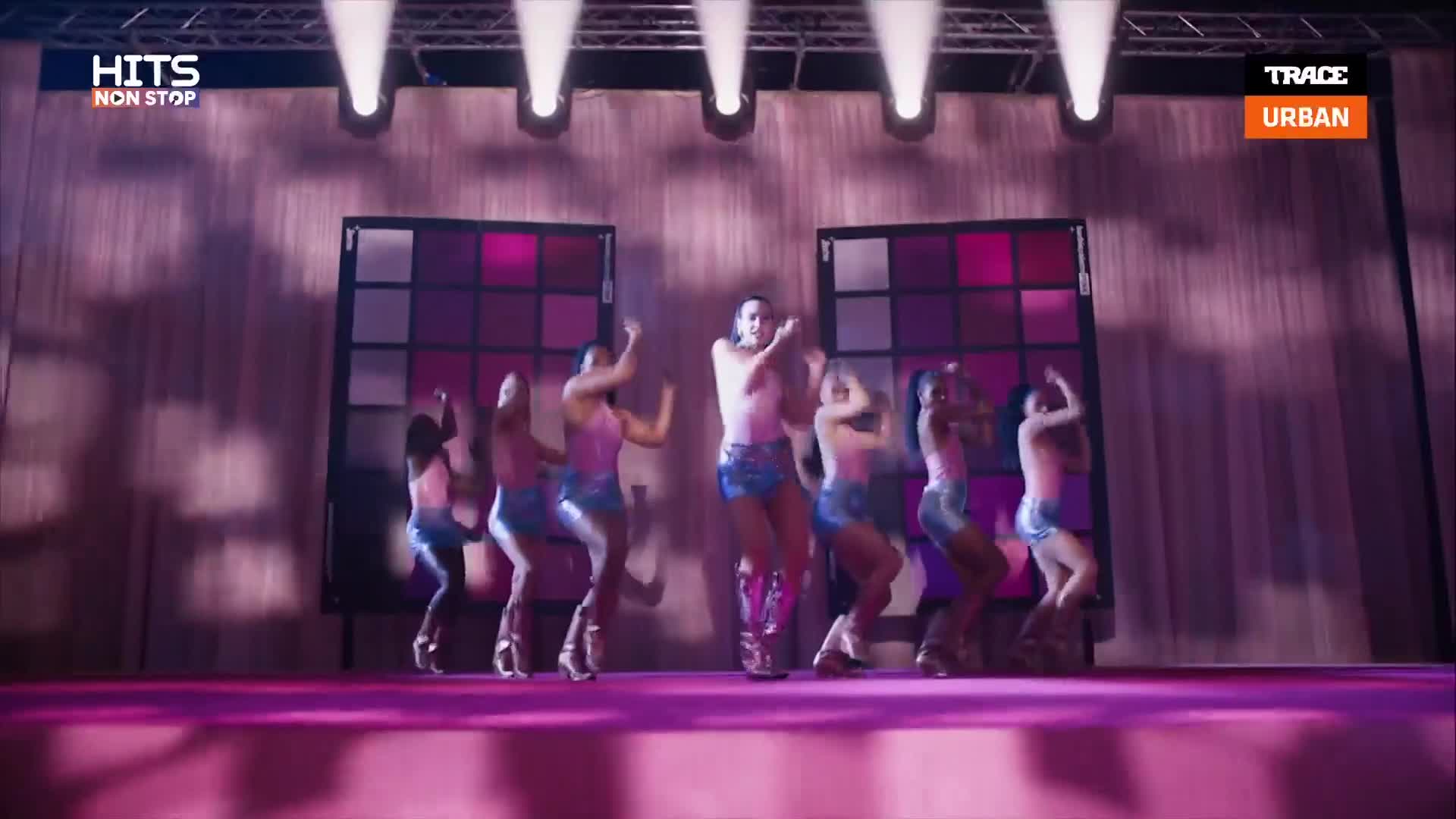 A group of dancers moves in unison on a stage, their arms raised. A central figure in sparkly boots leads the energetic choreography.