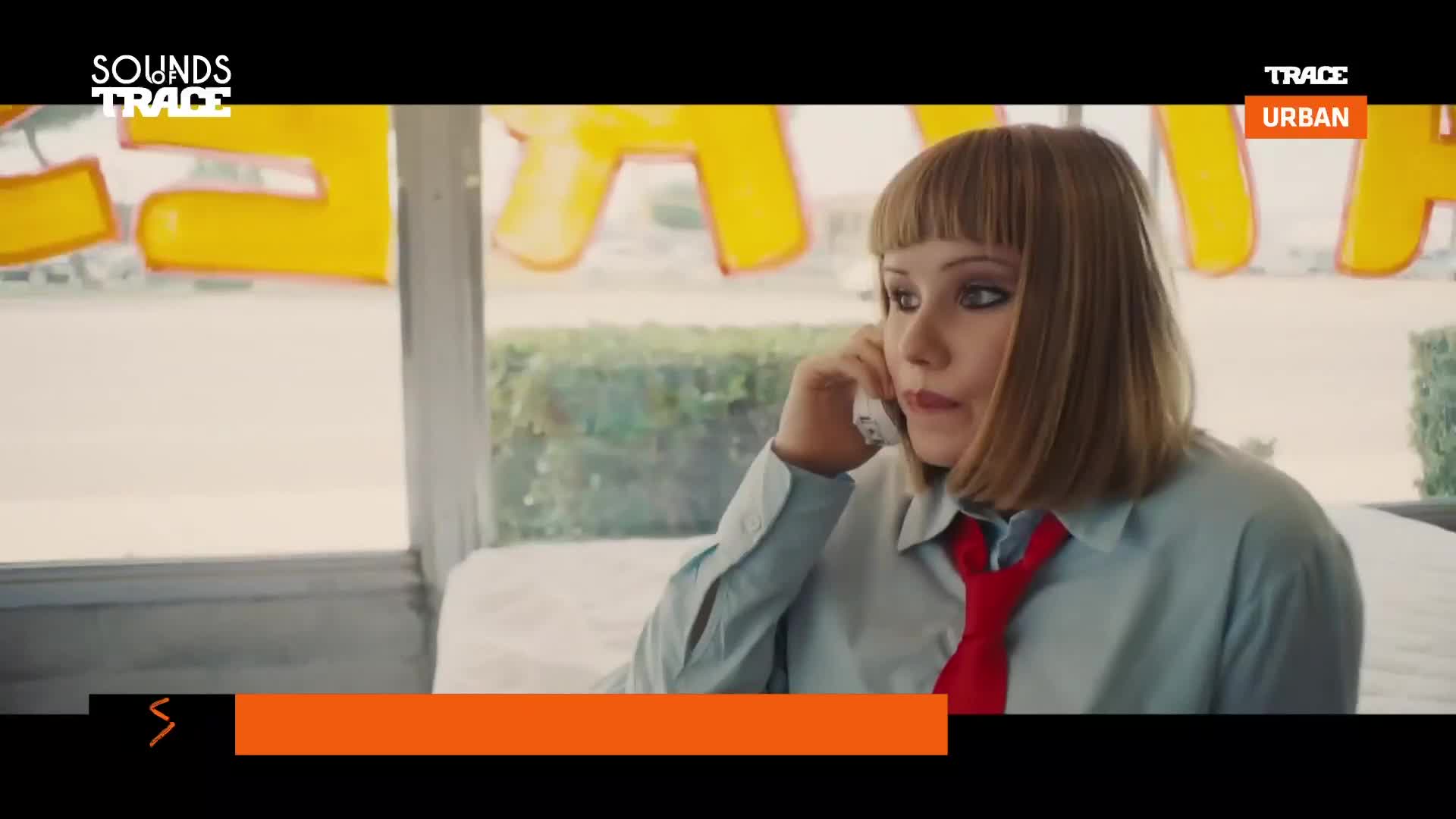 A young woman with a bright red tie speaks into a phone, her short blonde hair framing her face. Behind her, large yellow letters are visible through a window.