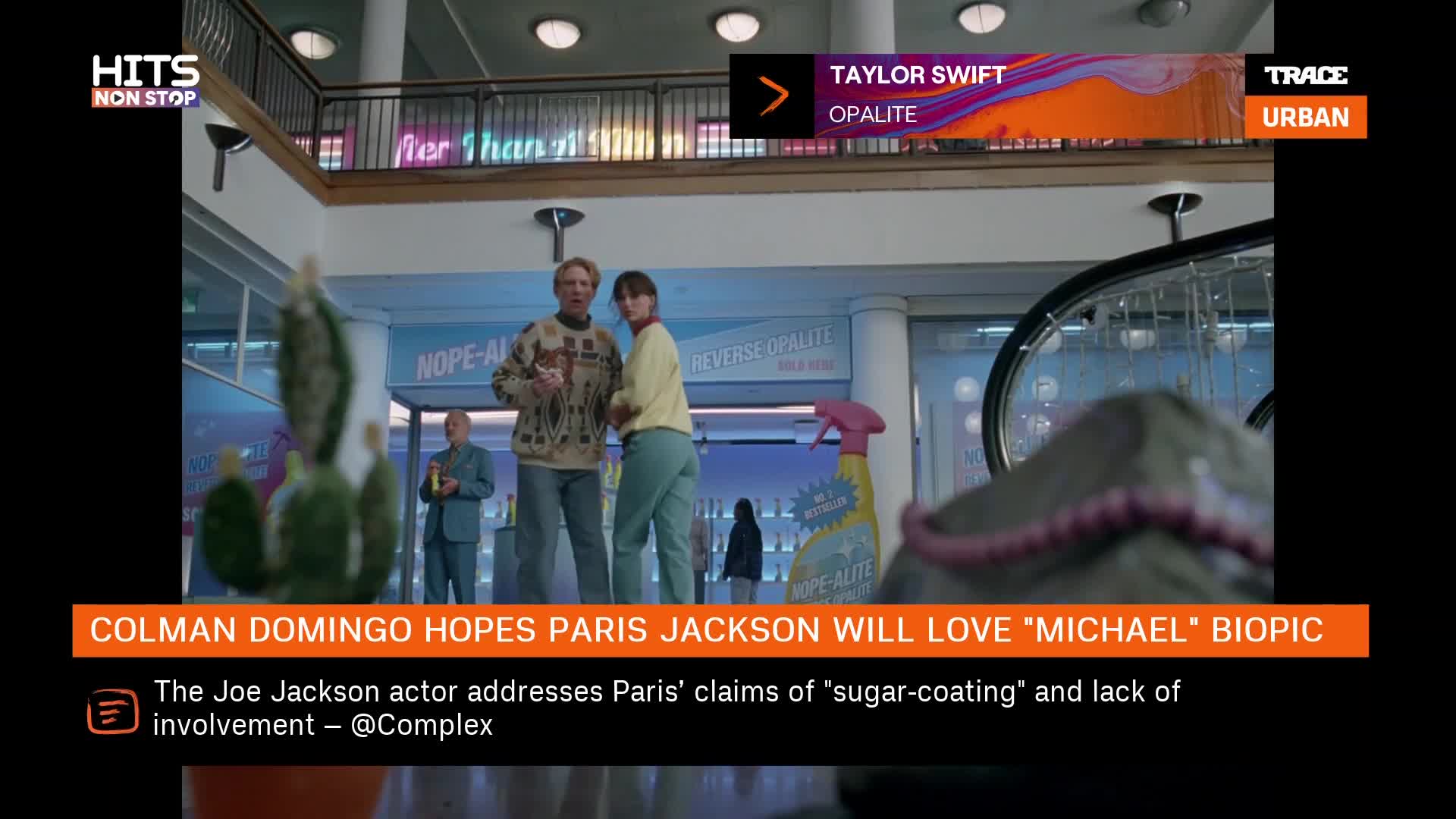 A man in a patterned sweater gestures animatedly while a woman in a yellow top stands beside him. Behind them, a pink spray bottle and cleaning supplies are displayed on a shelf.