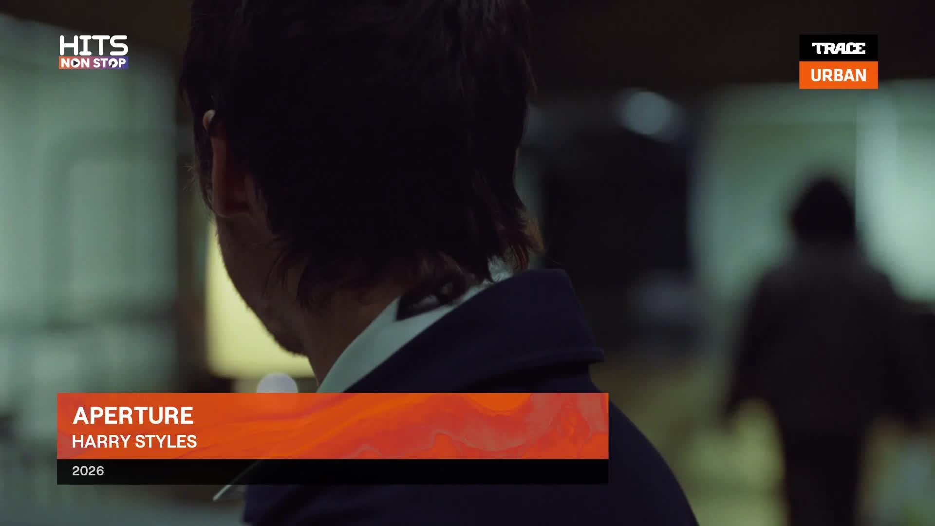 A man with dark hair turns away from the camera, his blue jacket collar visible. In the background, a shadowy figure walks away down a dimly lit hallway.