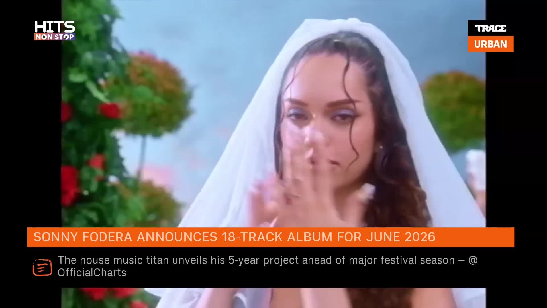 A woman in a white veil is captured in a close-up, her hands near her face. Behind her, lush green foliage with red accents creates a soft backdrop.