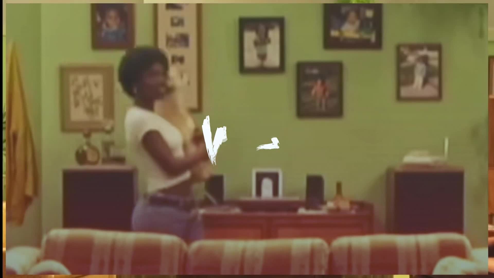 A young woman with an afro, wearing a white crop top and jeans, smiles as she holds a fluffy dog. Framed photos of athletes and family hang on the pale green wall behind her.