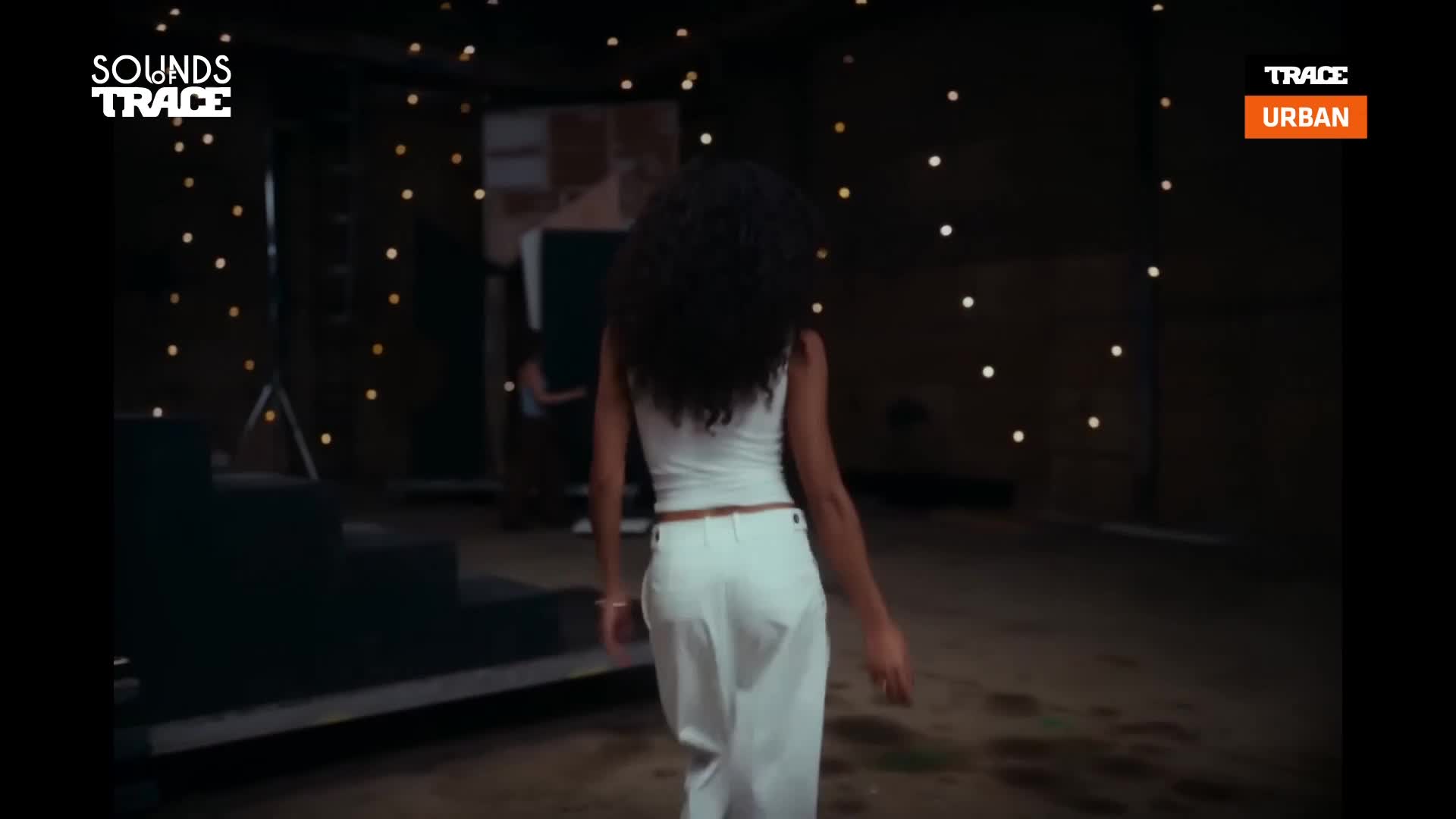 A woman in white pants and a tank top walks away from the camera, her dark curly hair flowing. The background is dimly lit with strings of small lights, hinting at a stage setup for a performance. A woman in white pants and a tank top walks away from the camera, her dark curly hair flowing. The background is dimly lit with strings of small lights, hinting at a stage setup for a performance.