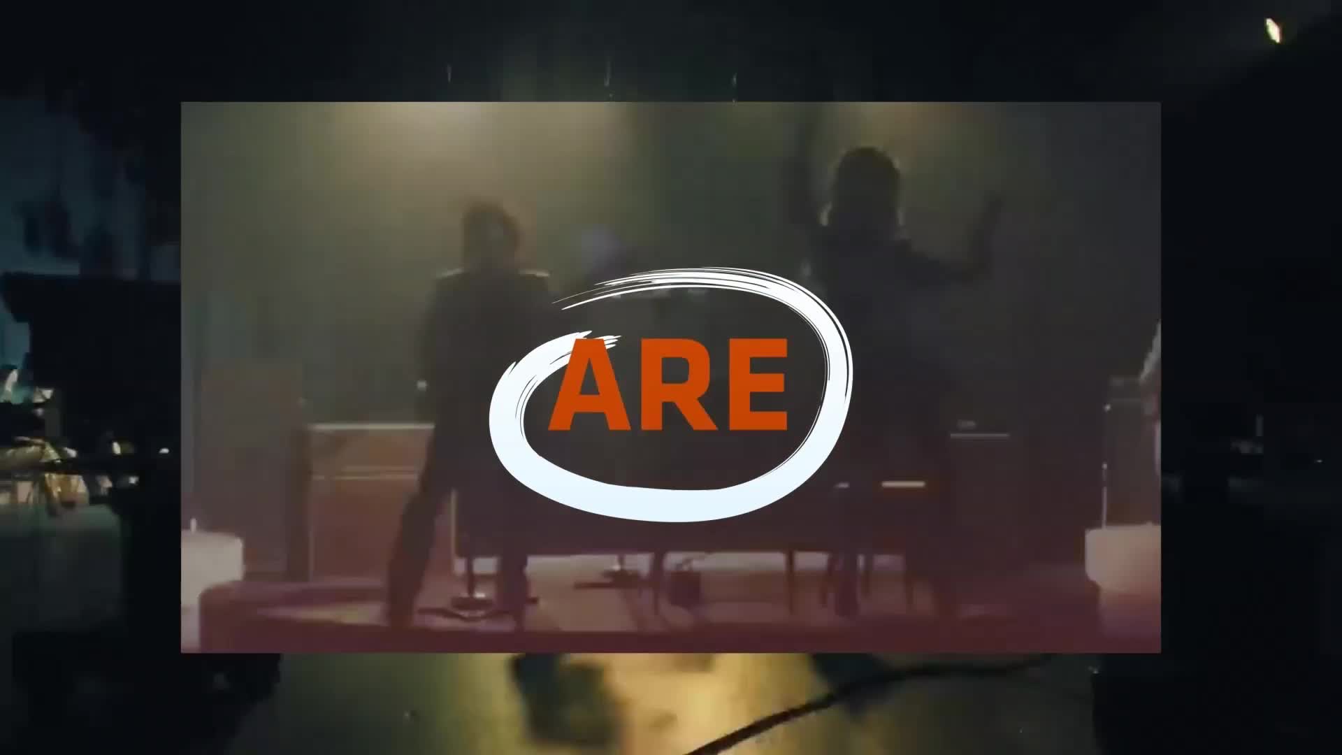 Two figures stand on a stage, one playing a keyboard. A white circle with the word "ARE" in orange overlays the scene. Two figures stand on a stage, one playing a keyboard. A white circle with the word "ARE" in orange overlays the scene.