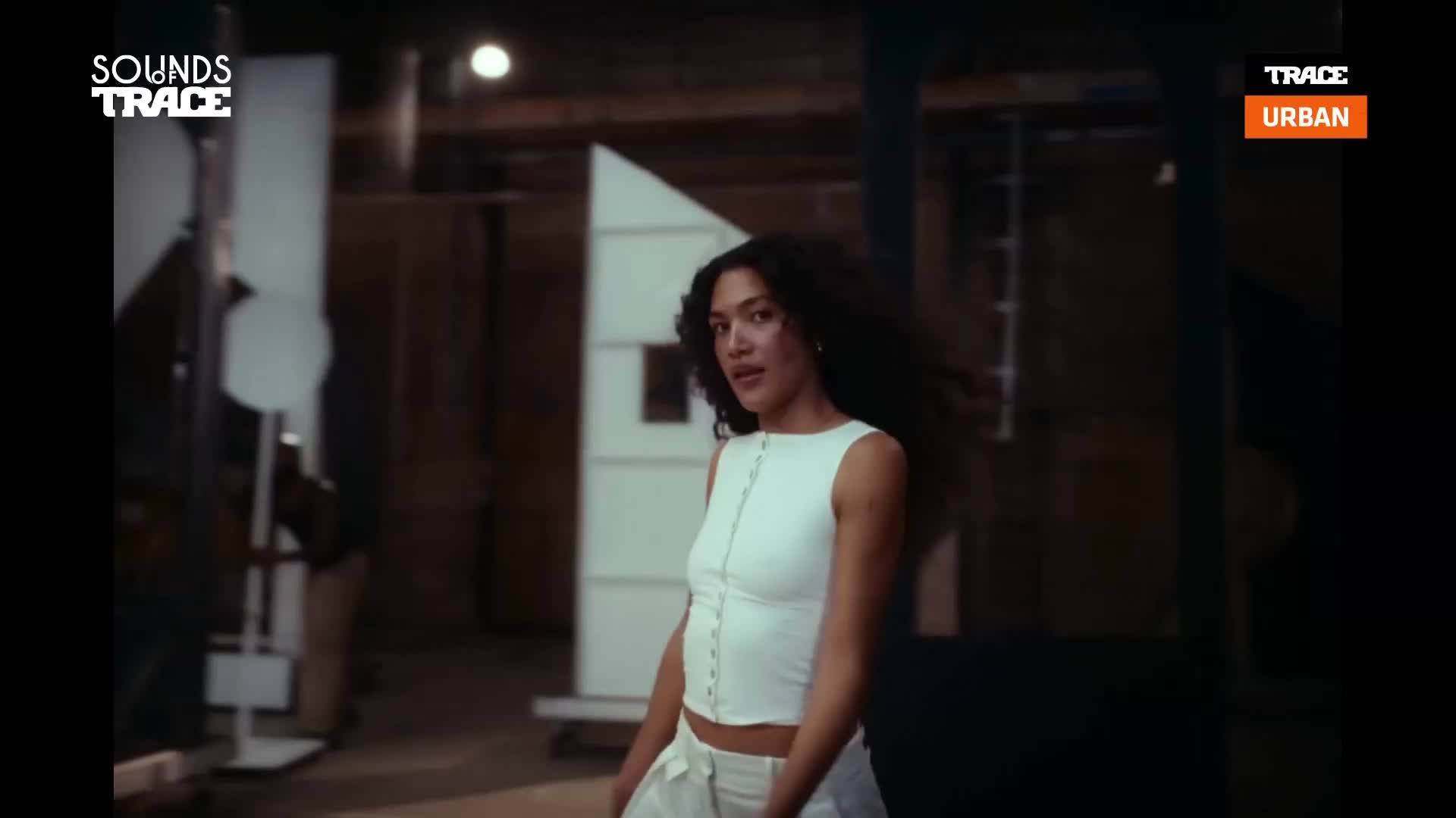 A young woman with dark, flowing hair turns her head, her white top catching the light. She stands in what looks like a workshop, with wooden structures and industrial beams around her.