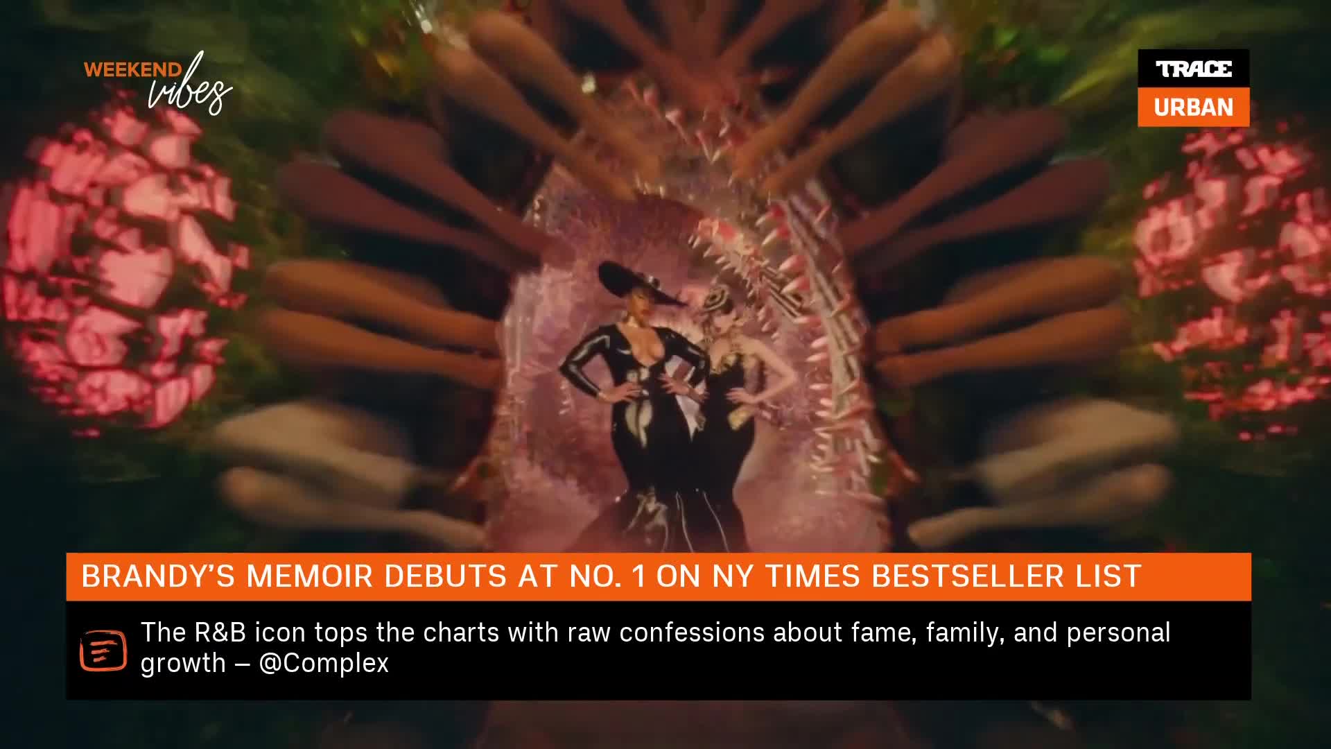 Two women in sleek, black outfits stand center stage, framed by a surreal, organic-looking structure. The Trace Urban logo is visible in the corner, hinting at a music video or performance.