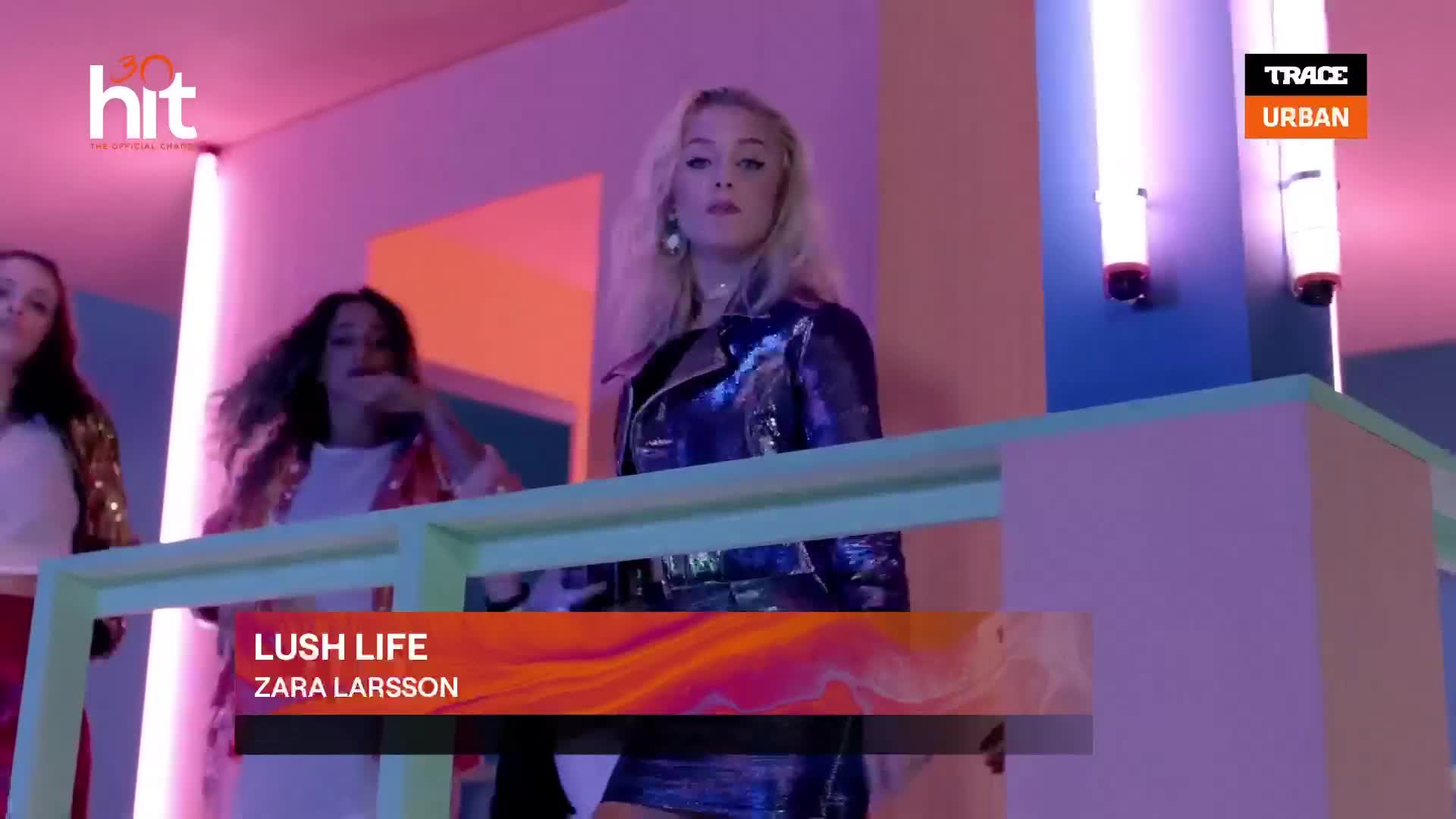 A blonde woman in a shiny jacket stands center, looking directly ahead. Behind her, two other women are visible, one with curly dark hair, all in a brightly lit, modern space.