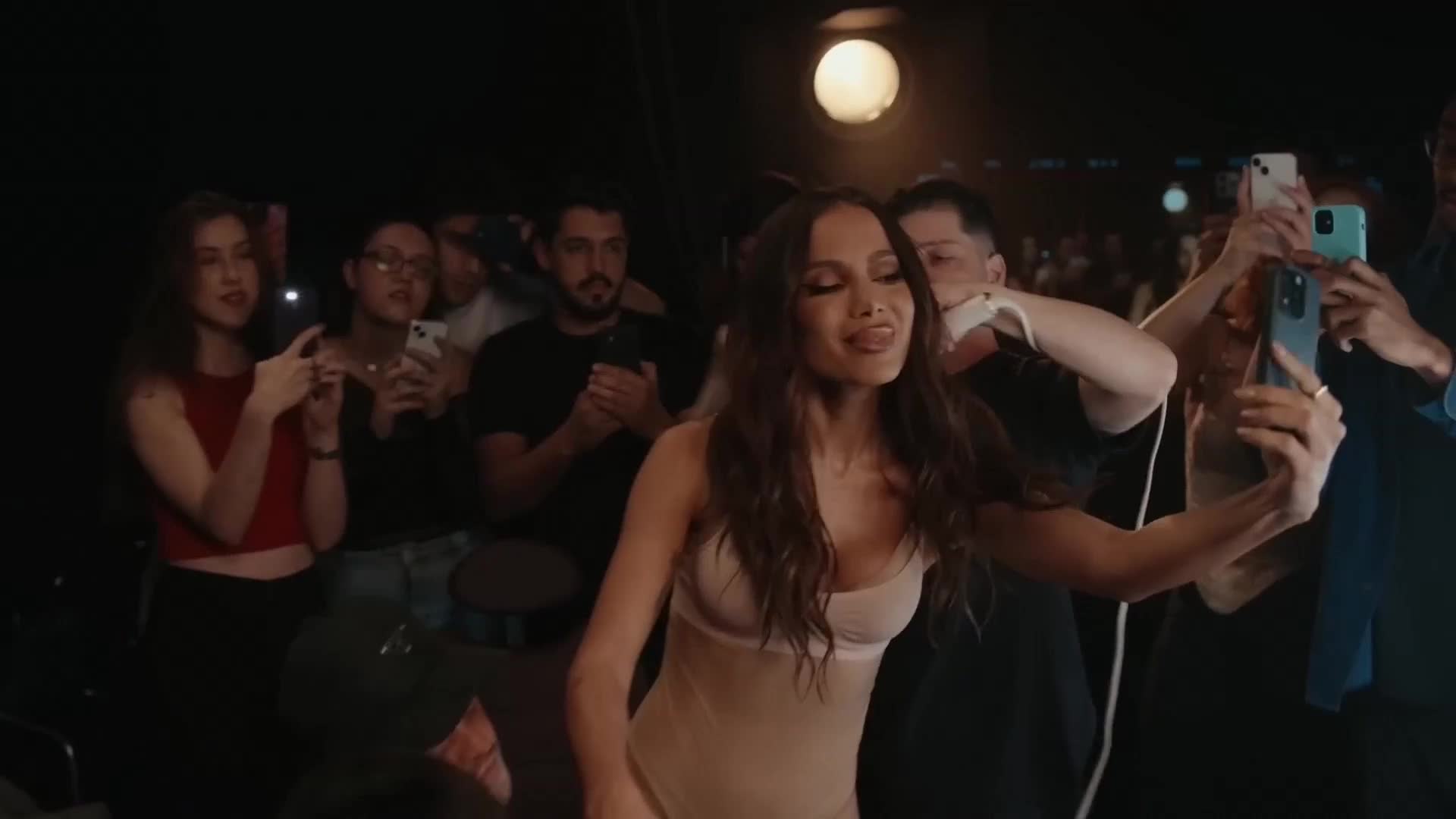 A singer in a light-colored leotard sticks out her tongue while posing for selfies. The crowd around her, likely at a concert in France, holds up their phones, capturing the moment.
