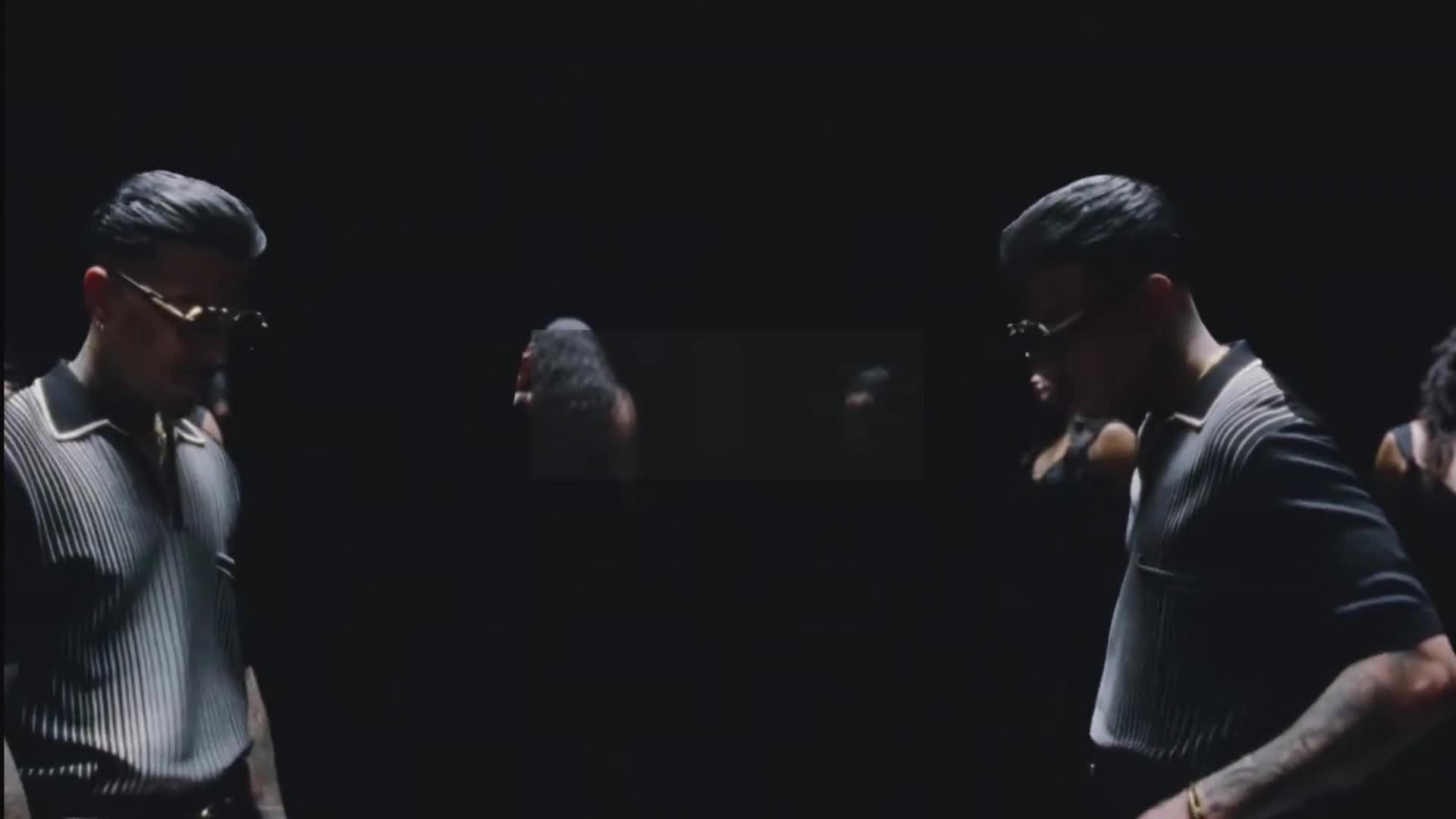 Two men in stylish polos and sunglasses move in unison, their reflections mirroring them in the darkness. Behind them, shadowy figures with long hair appear and disappear.