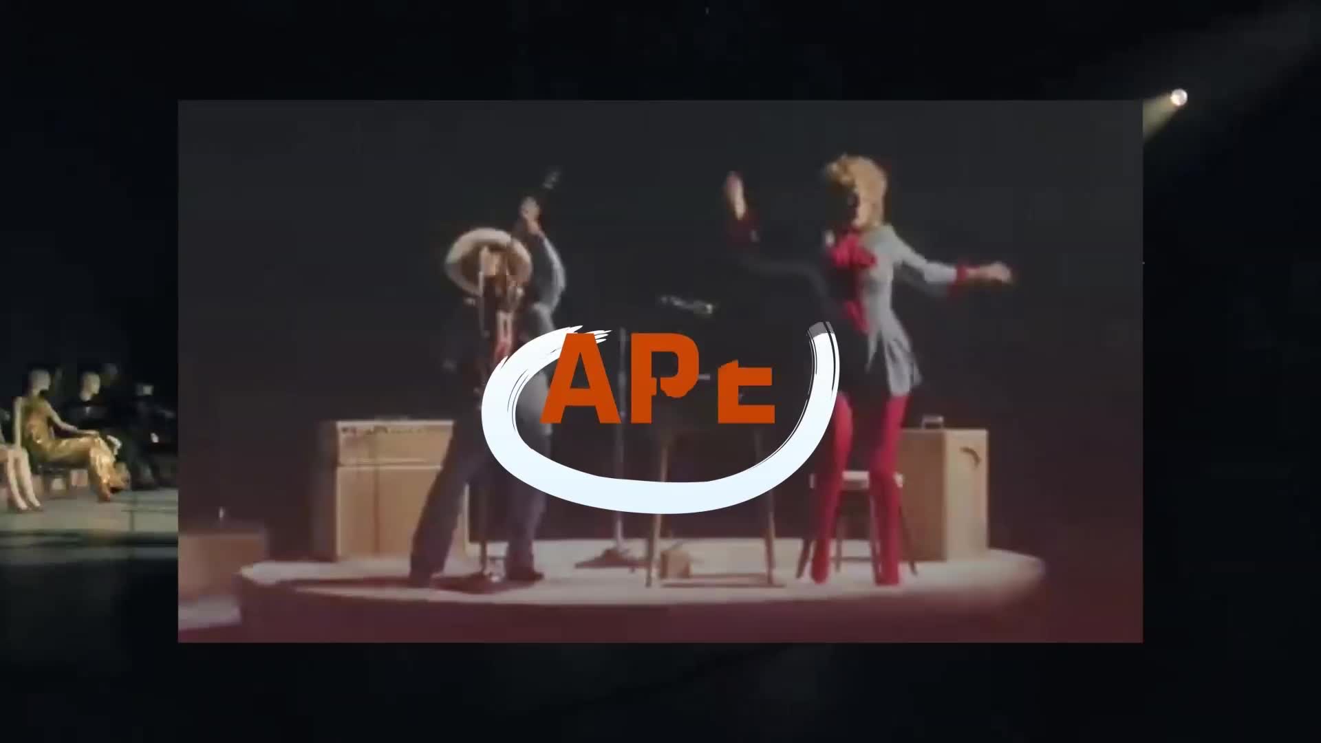 A woman in red tights and a grey tunic dances energetically on stage. Beside her, a man in a wide-brimmed hat plays a guitar.