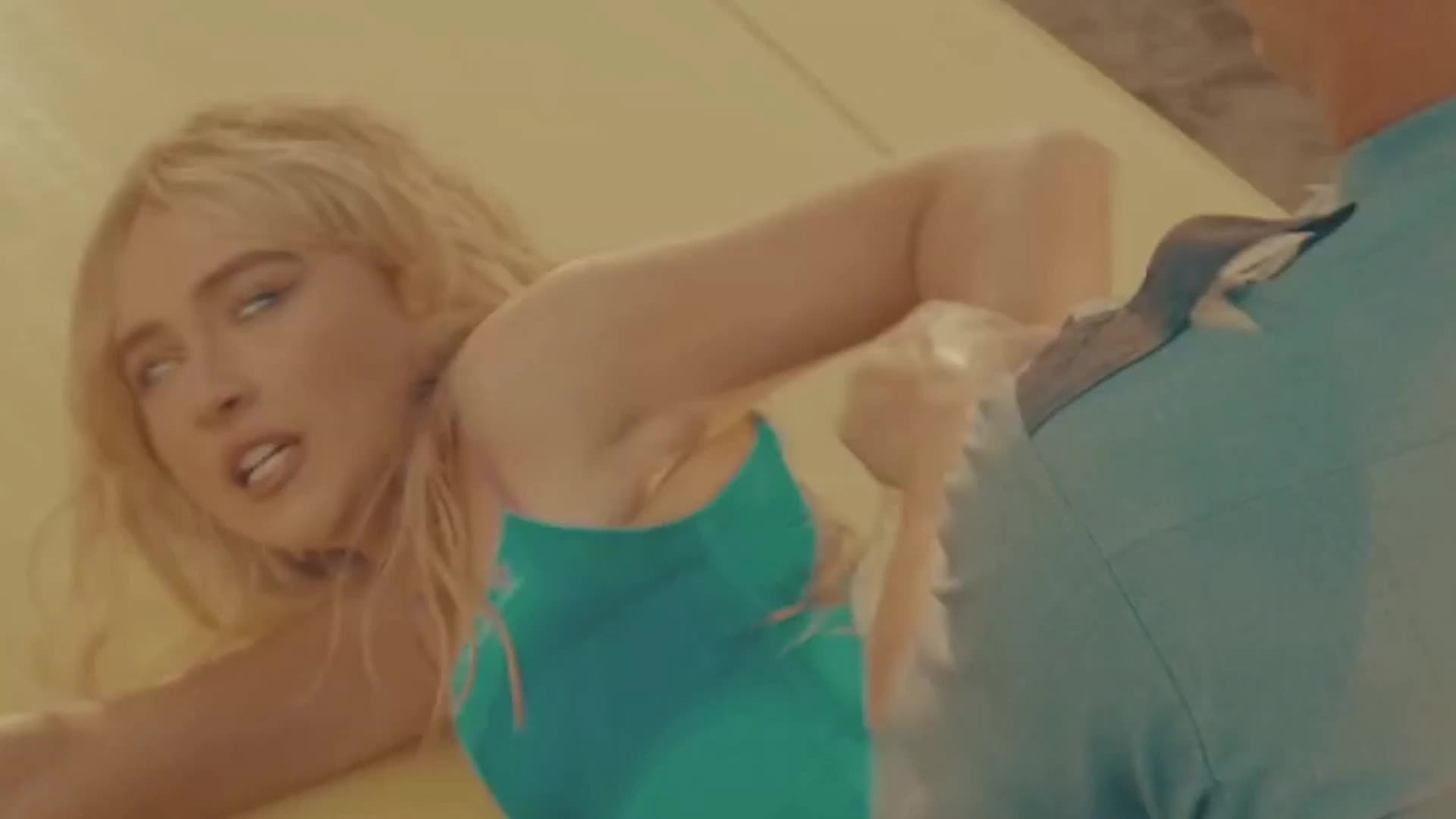 A blonde woman in a teal swimsuit lunges forward, her arm outstretched. A man in a light blue shirt turns away as she reaches for him.