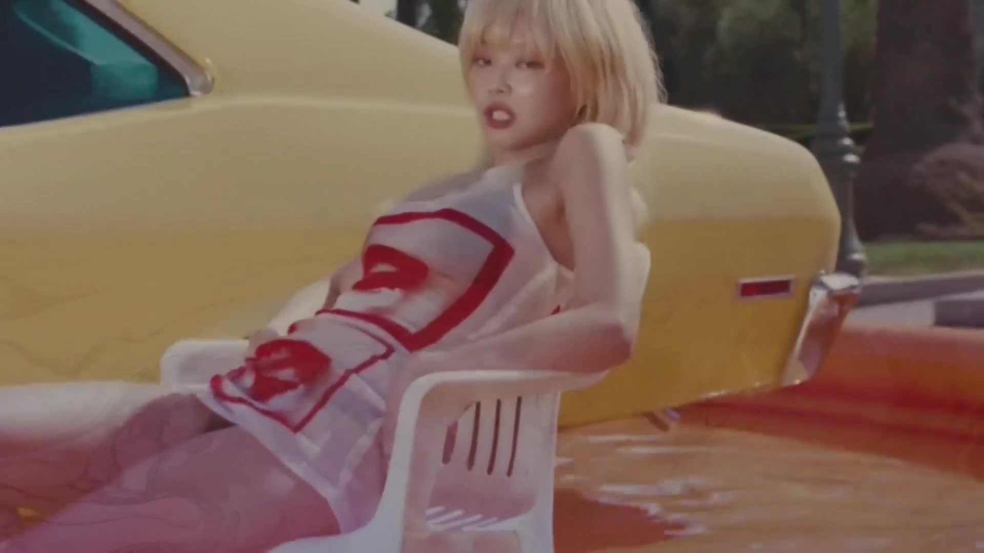 A woman with blonde hair reclines in a white chair, her sheer dress patterned with red lips. Beside her, a bright yellow car curves into the frame.