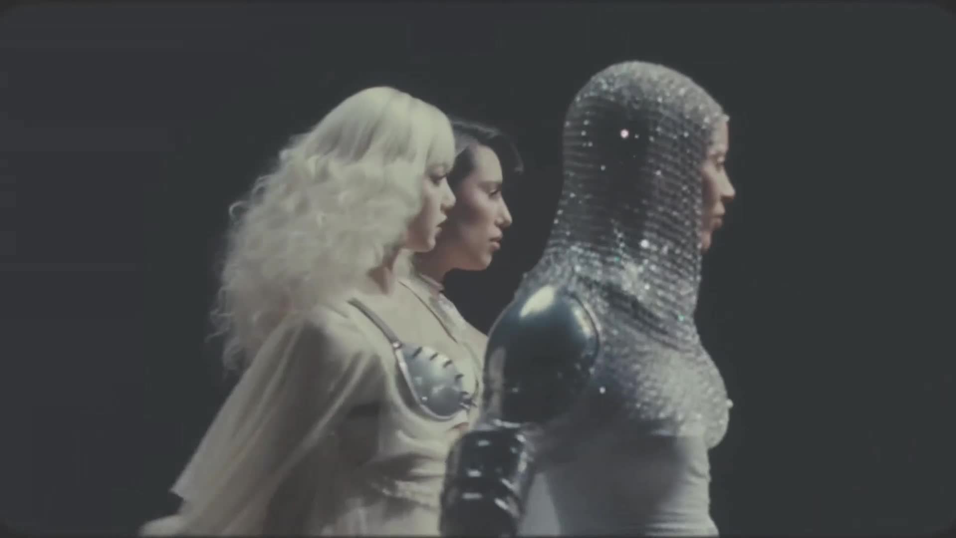 A figure in a shimmering chainmail hood and metallic armor walks forward, followed by two women in avant-garde outfits. The scene has the stylized, futuristic feel of a Trace Urban production.