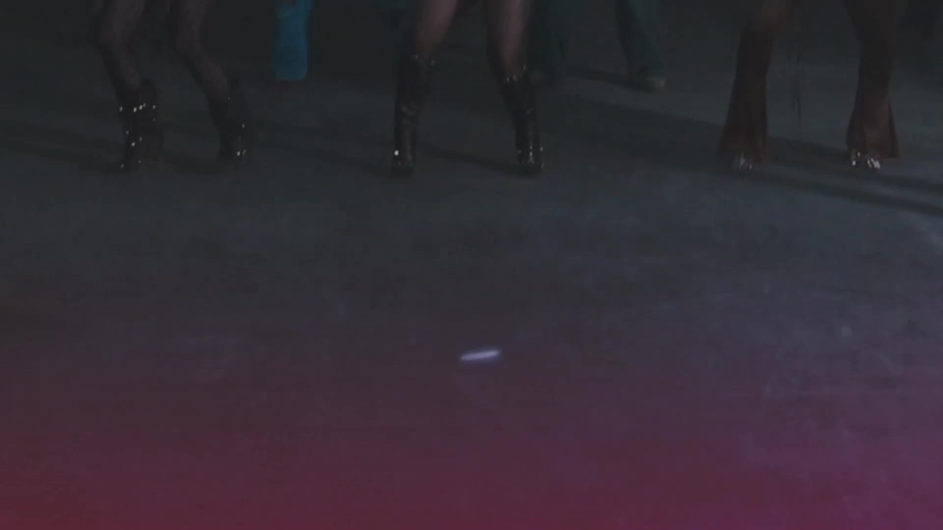 A thin, glowing line streaks across the dark floor as dancers in flashy boots move above. The stage lights cast a purple haze, hinting at a lively performance.