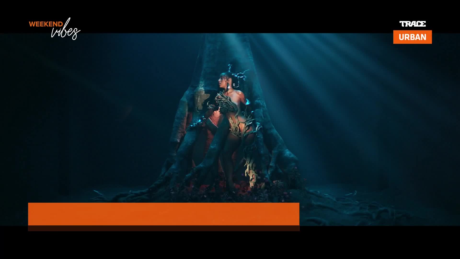 Sunlight streams down through dark water, illuminating a woman adorned with leaves and vines. She sits within the gnarled roots of a massive tree, its branches twisting around her.