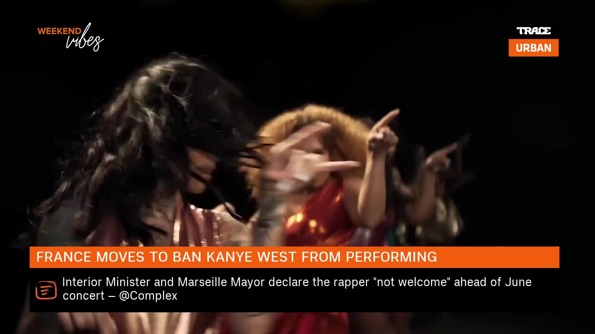 Dancers move their arms in unison, their fingers pointing towards the audience. The news ticker below announces France's decision to ban Kanye West from performing.