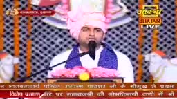 A man in a pink turban and blue scarf speaks into a microphone, likely for the Aastha Bhajan program broadcast from India. The backdrop features a patterned design and a banner with Hindi script.

