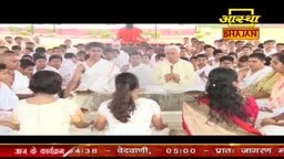 A large group of people in India are gathered, with some in white clothing, listening to a man in a yellow shirt. The Aastha Bhajan logo is visible, indicating a religious broadcast.
