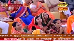 People in colorful saris and headscarves are seated, many looking towards something out of frame. The "Aastha Bhajan LIVE" logo is visible, suggesting a live broadcast of a religious event.
