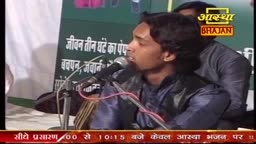 A young man in a dark shirt sings into a microphone, his face illuminated by the stage lights. Behind him, the logo for Aastha Bhajan is visible, and a man in a similar shirt is partially visible.
