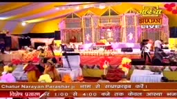 A stage is set with a backdrop that reads "Aastha Bhajan Live." People are gathered, some wearing turbans, as a broadcast is underway in India.
