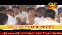 A group of people, many wearing white, stand close together, their hands clasped in prayer. The Aastha Bhajan logo and Hindi text appear on a screen below.
