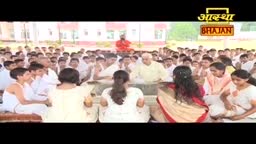 People in white and cream clothing sit in rows, facing a central area where a few figures are seated. A man in a red robe stands in the background, and the "Aastha Bhajan" logo is visible at the top.
